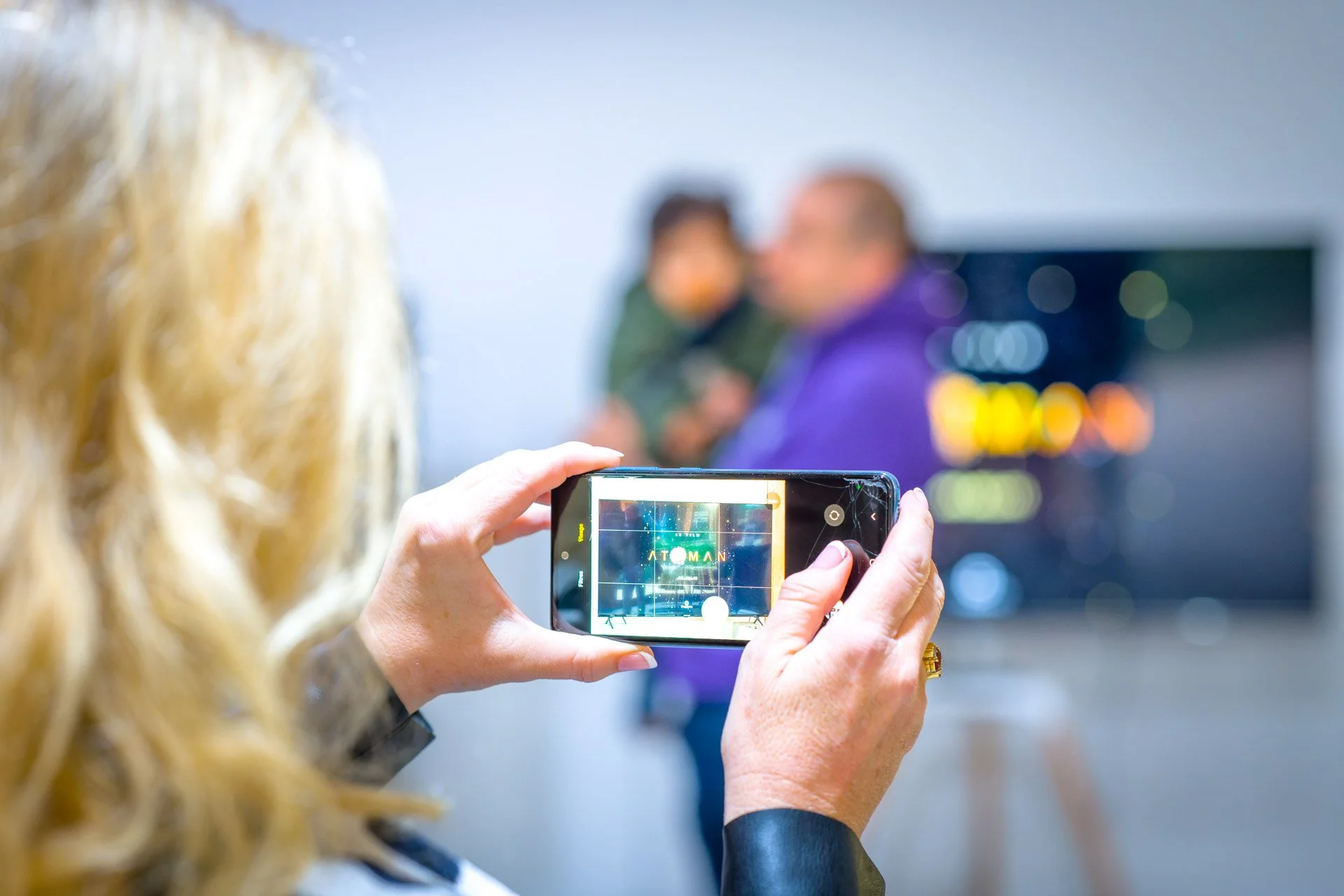 Personne tenant un smartphone prenant une photo, avec deux personnes floues à l'arrière-plan.