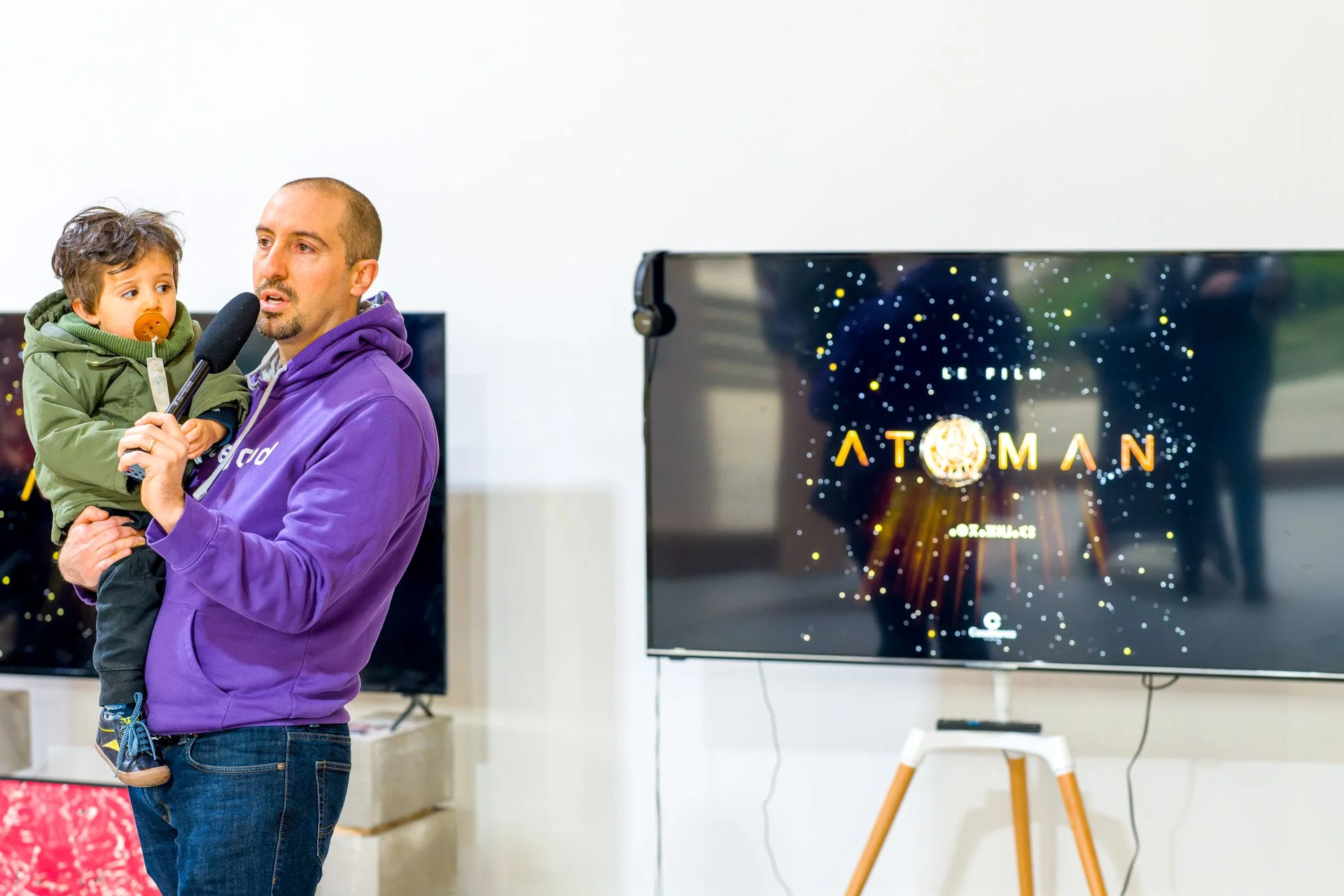 Un homme portant un enfant parle dans un micro devant un écran affichant "Le film Atoman".