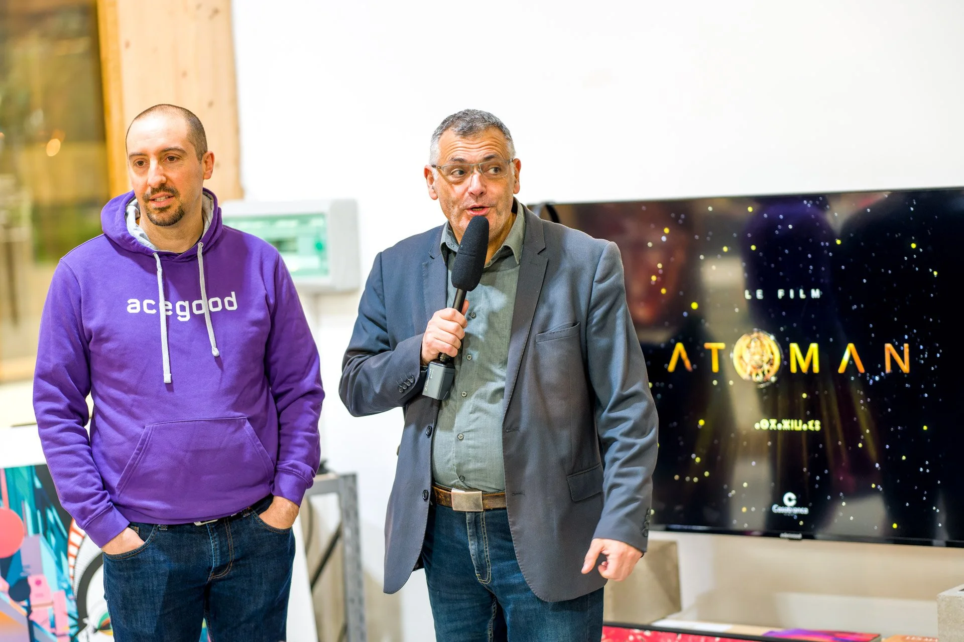 Deux hommes lors d'une conférence, l'un en pull violet "acegood", l'autre en costume tenant un micro. En arrière-plan, écran affichant "Le film ATOMAN". Environnement intérieur lumineux.