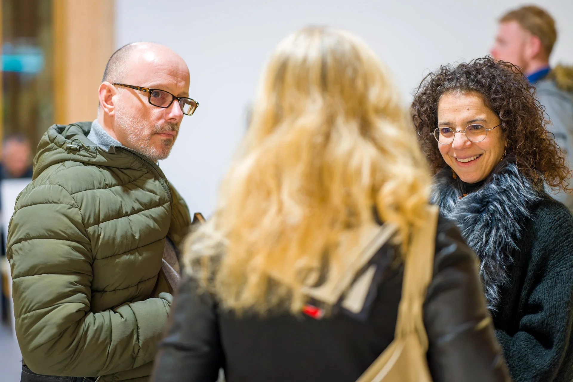 Plusieurs personnes discutent dans un environnement intérieur. Un homme porte une veste verte et des lunettes, et une femme sourit, portant une écharpe en fourrure.