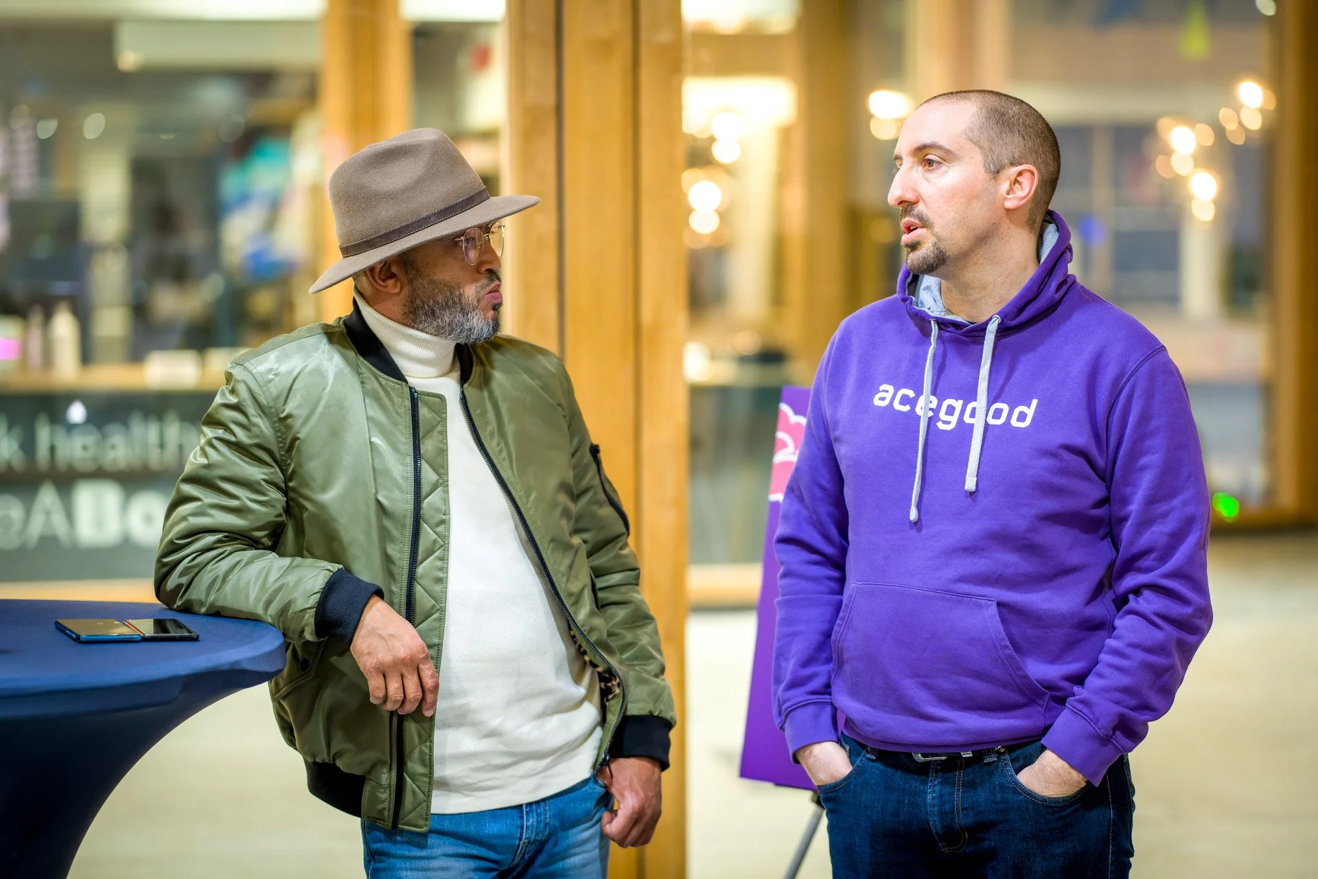 Deux hommes discutent, l'un portant un chapeau et une veste verte, l'autre un sweat violet avec l'inscription 'acegood', à l'intérieur d'un bâtiment moderne.
