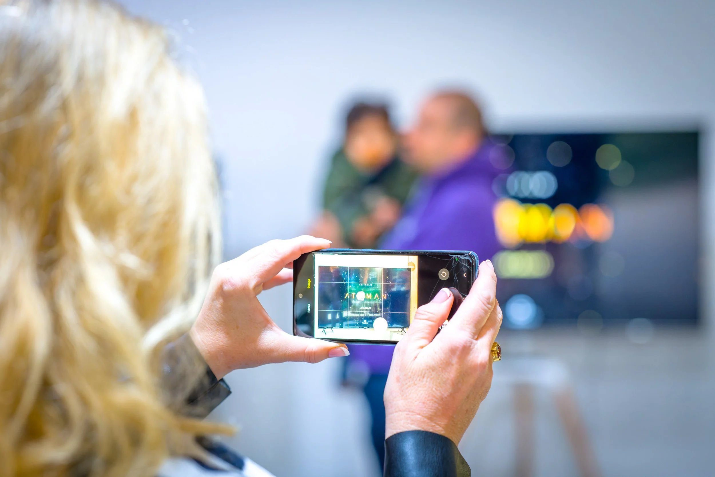 Une personne avec des cheveux blonds qui prend une photo avec son téléphone d'un homme qui tient un enfant dans un environnement intérieur.