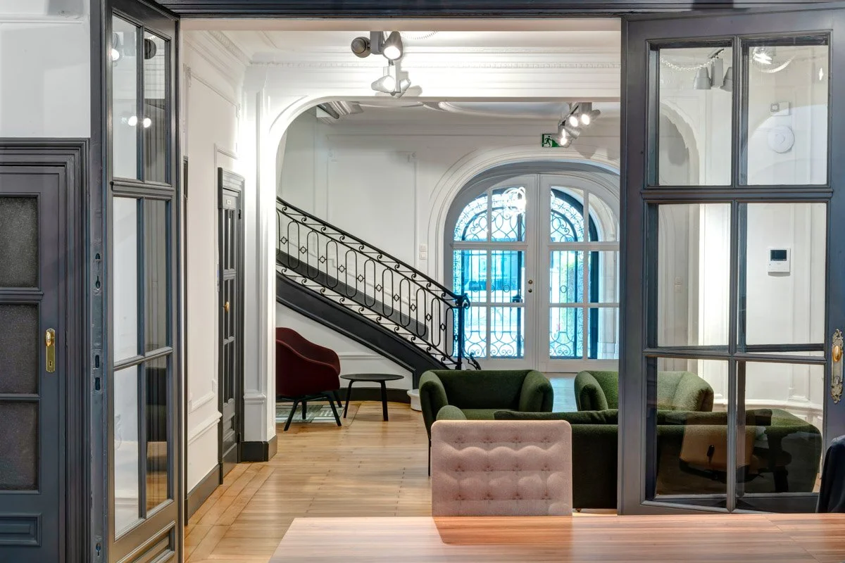 Un salon avec fauteuils, visible à travers une cloison vitrée, avec un escalier et une grande fenêtre cintrée à l’arrière-plan.