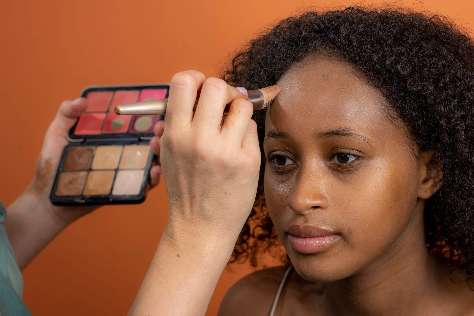 Une personne applique du maquillage sur le front d'une femme à l'aide d'un pinceau, avec une palette de maquillage visible.