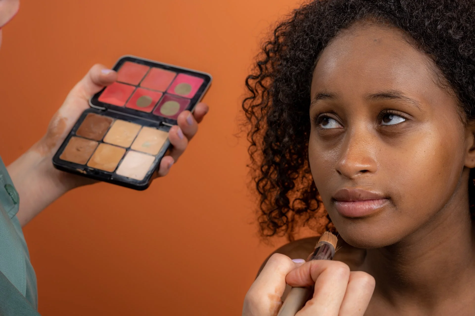 Jeune femme recevant un maquillage avec une palette de fards à paupières, fond orange.