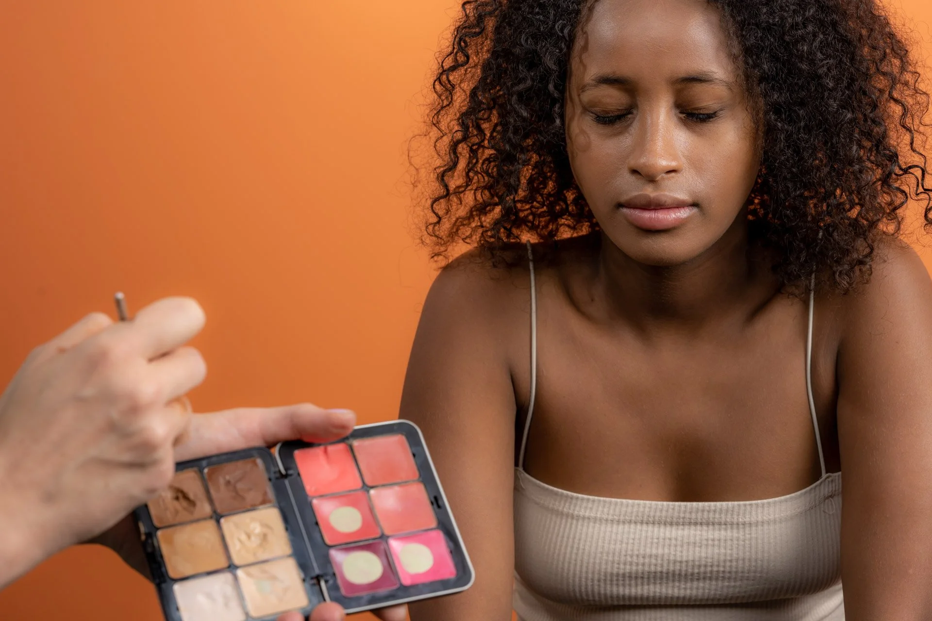 Une femme aux cheveux bouclés se fait maquiller, les yeux fermés, avec une palette de maquillage à l'avant-plan.