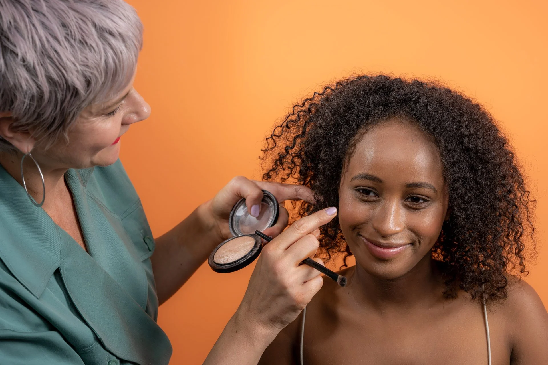 Une femme applique du maquillage sur une autre femme souriante avec un fond orange.