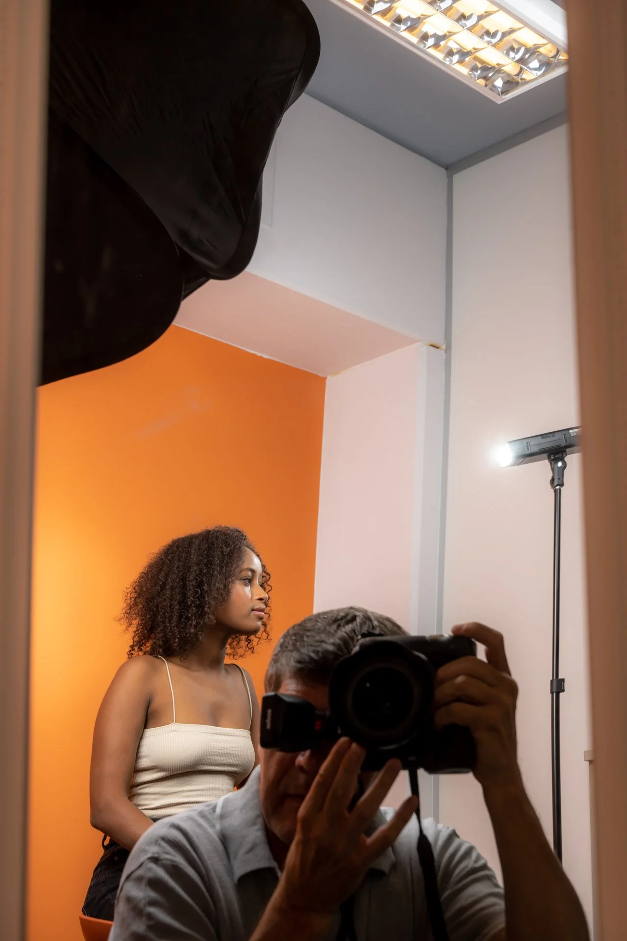 Dans le reflet d’un miroir, un photographe prend une photo d’une femme assise en studio devant un fond orange.