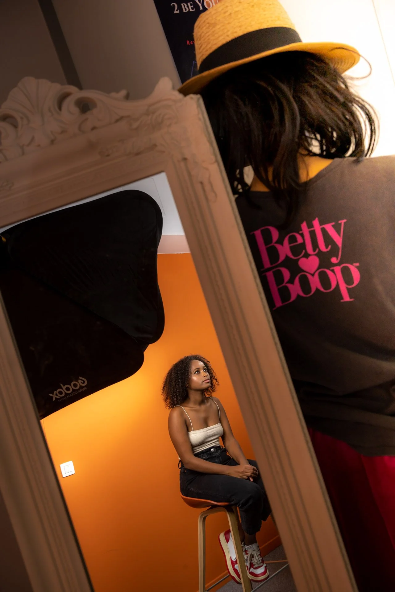 Un miroir reflète une femme assise sur un tabouret devant un mur orange, un réflecteur noir à côté. Le reflet montre une personne portant un chapeau de paille et un t-shirt "Betty Boop".