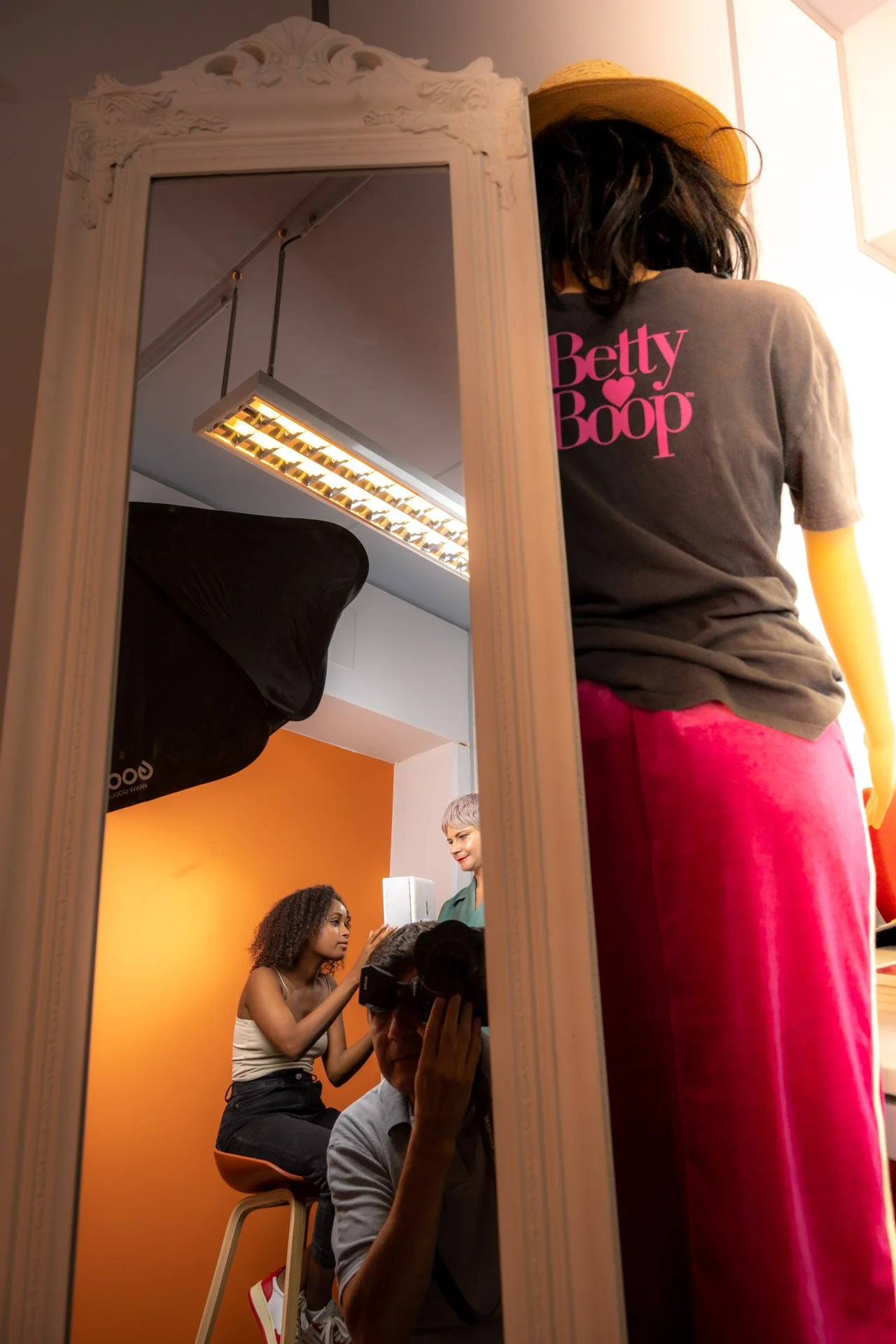 Une personne portant un chapeau, un tee-shirt Betty Boop et un pantalon rose se tient devant un miroir reflétant une séance photo en studio.