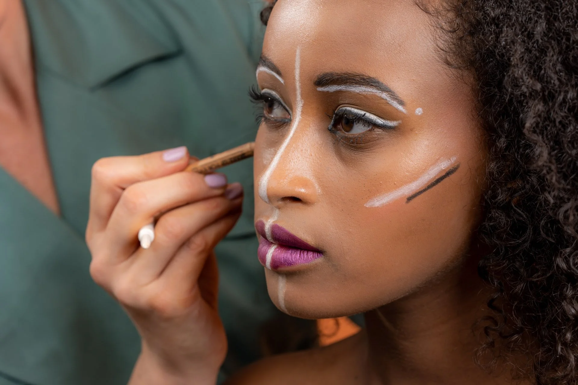 Personne appliquant des motifs de maquillage artistique blanc et noir sur une femme au teint foncé.
