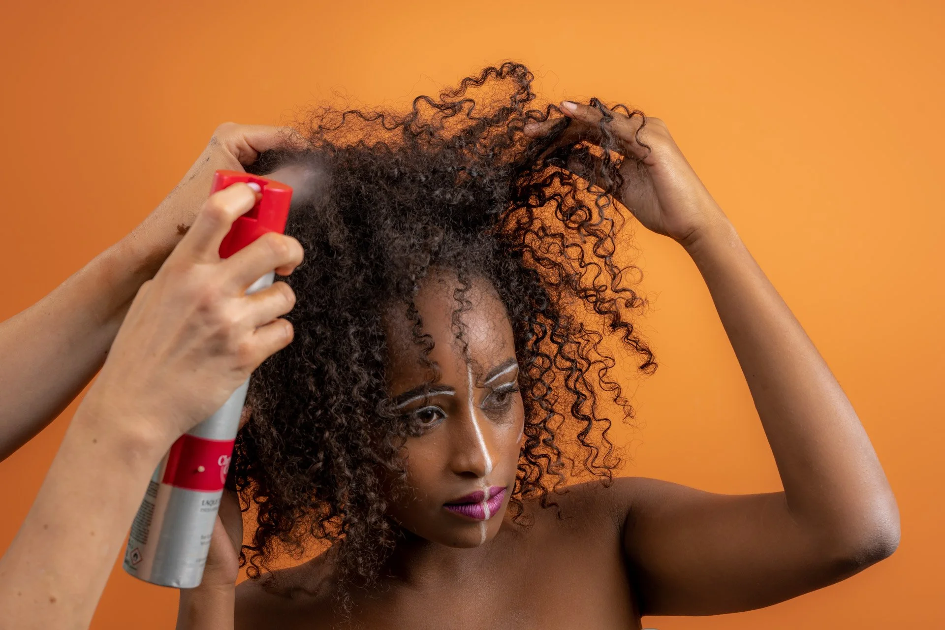 Une personne aux cheveux bouclés se faisant vaporiser de la laque sur les cheveux par une autre personne, sur un fond orange.