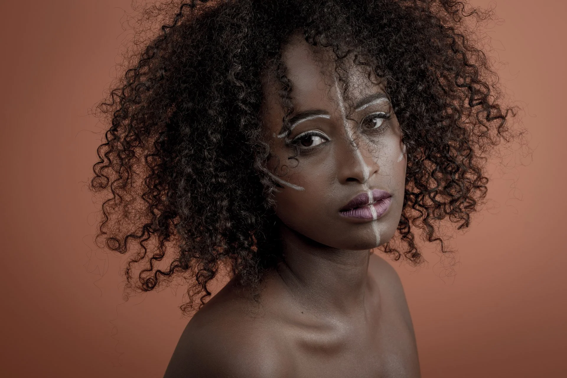 Portrait d'une femme à la peau foncée avec des cheveux bouclés, sur fond orange, maquillage artistique blanc sur le visage.