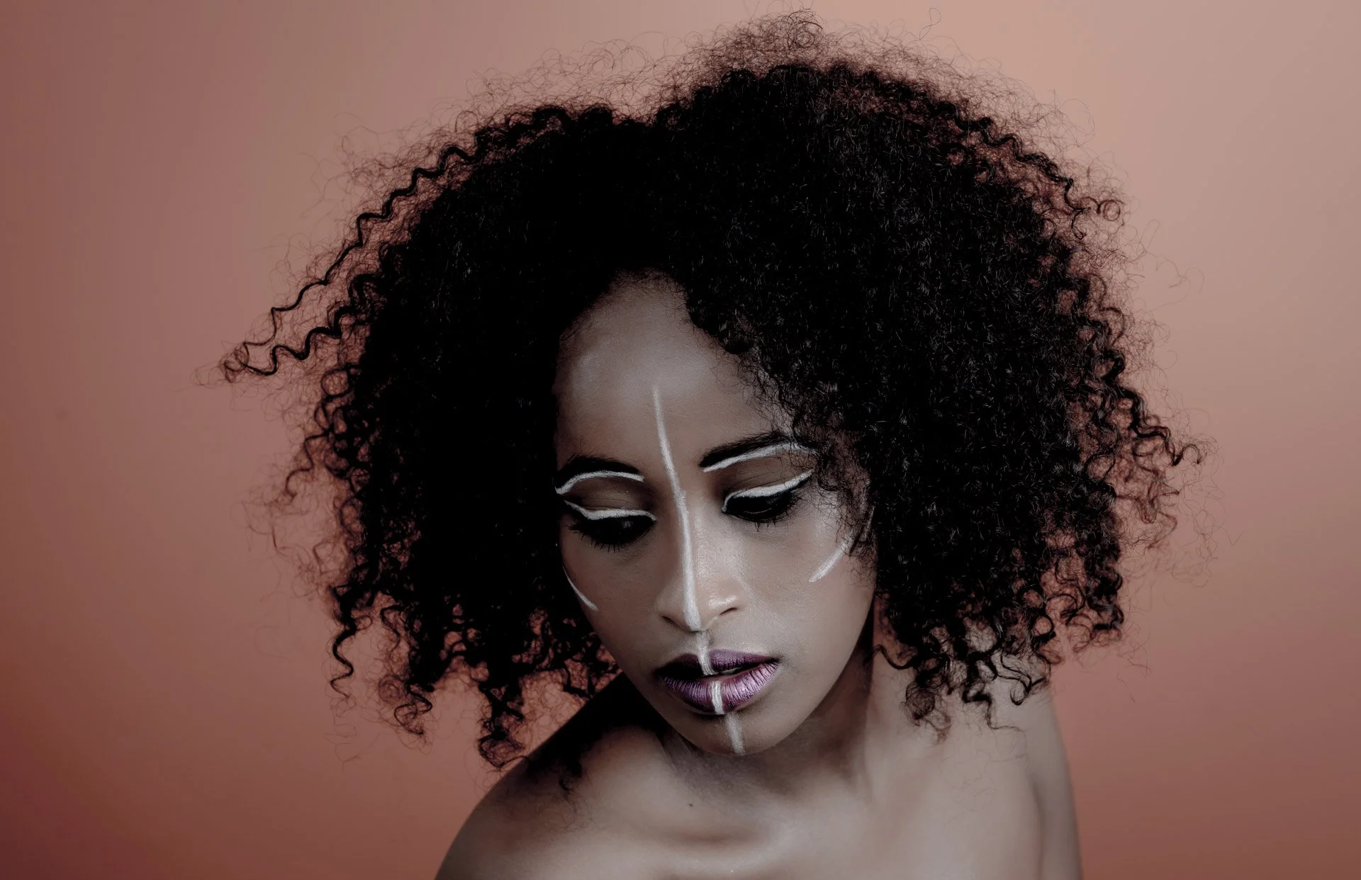Une personne avec des cheveux bouclés noirs et des motifs de maquillage blancs sur le visage sur fond orange.