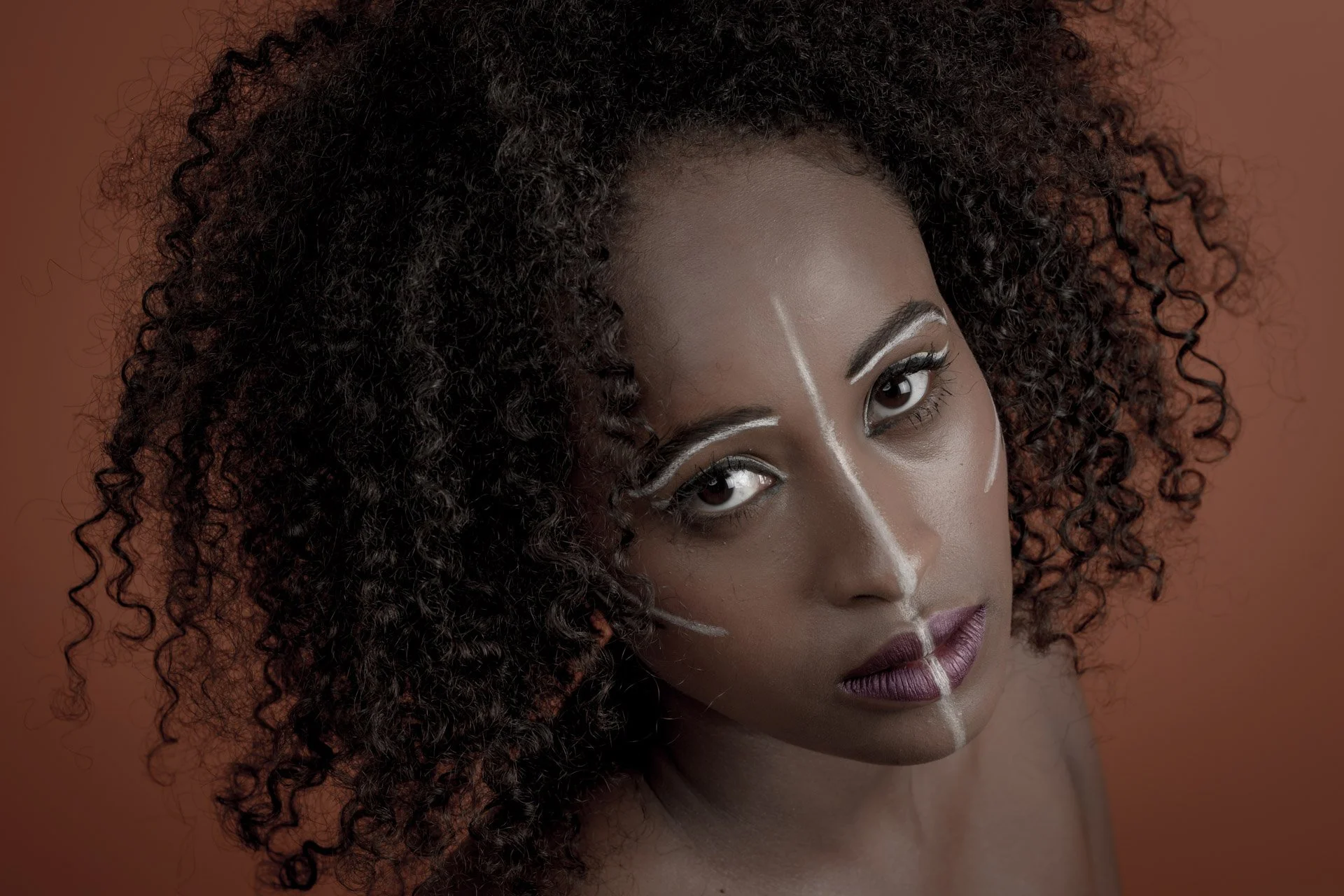 Femme avec maquillage artistique, peintures blanches sur le visage, cheveux bouclés, fond brun.
