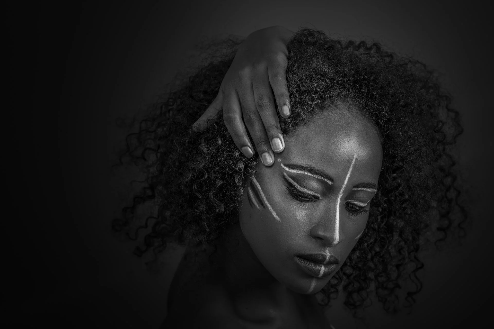 Portrait en noir et blanc d'une femme avec des motifs de peinture blanche sur le visage et une main tenant ses cheveux bouclés.