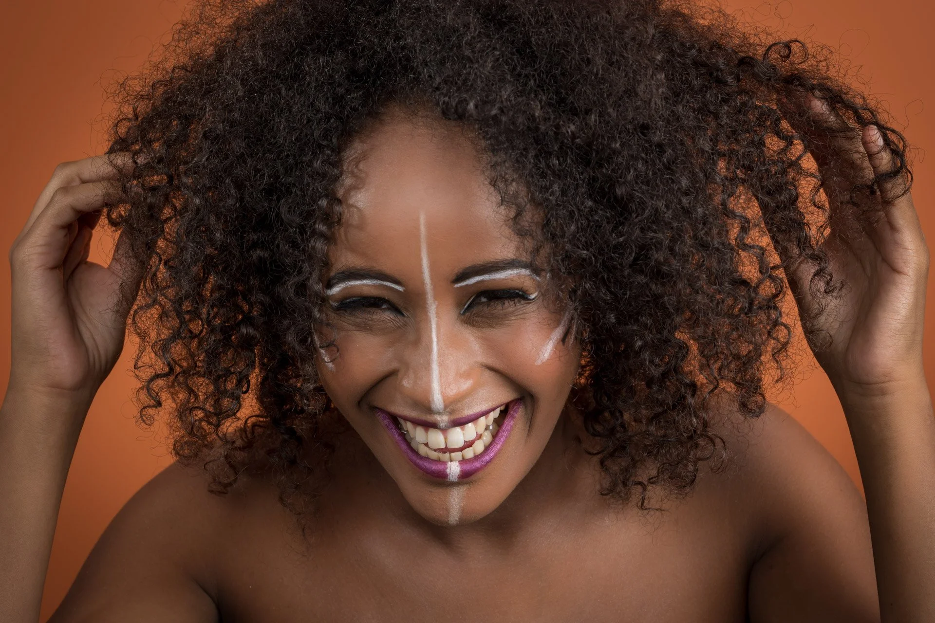Femme souriante avec des cheveux bouclés et maquillage artistique sur fond orange.