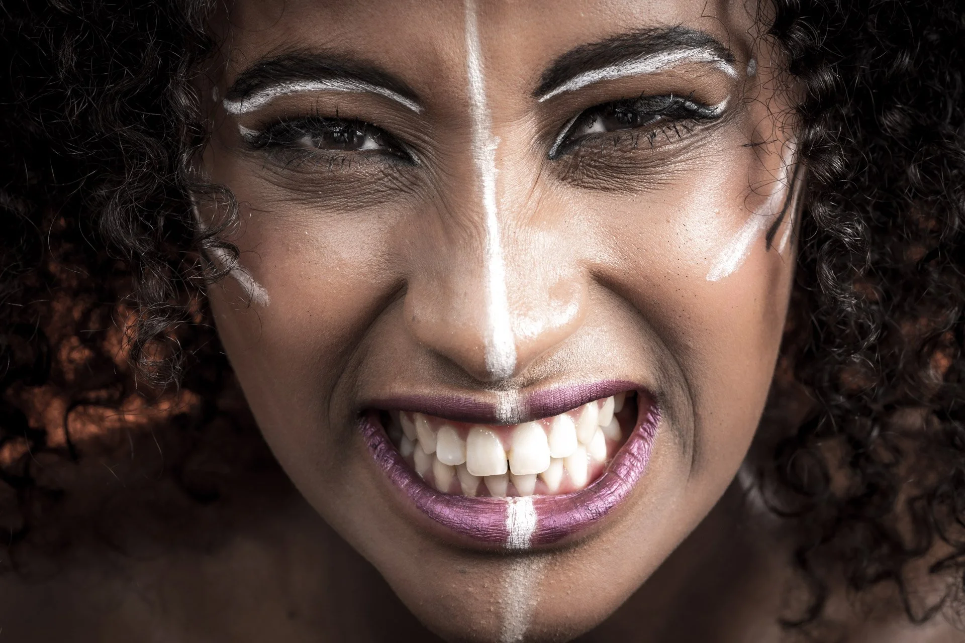 Gros plan d'une personne avec du maquillage tribal et des cheveux bouclés, montrant ses dents.