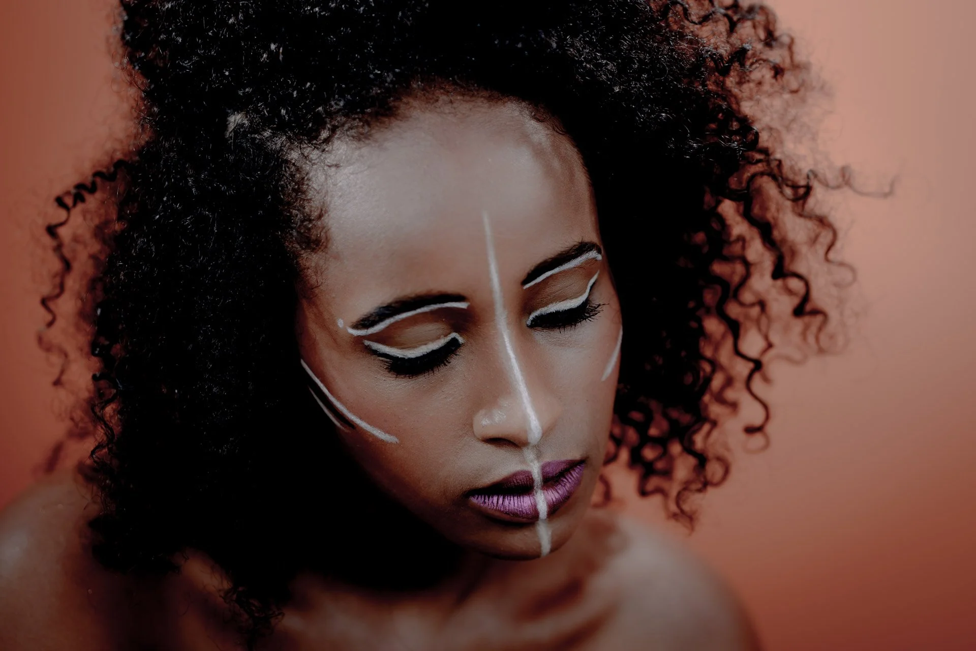 Femme avec des cheveux bouclés, maquillage artistique avec des lignes blanches sur le visage, sur fond orange.