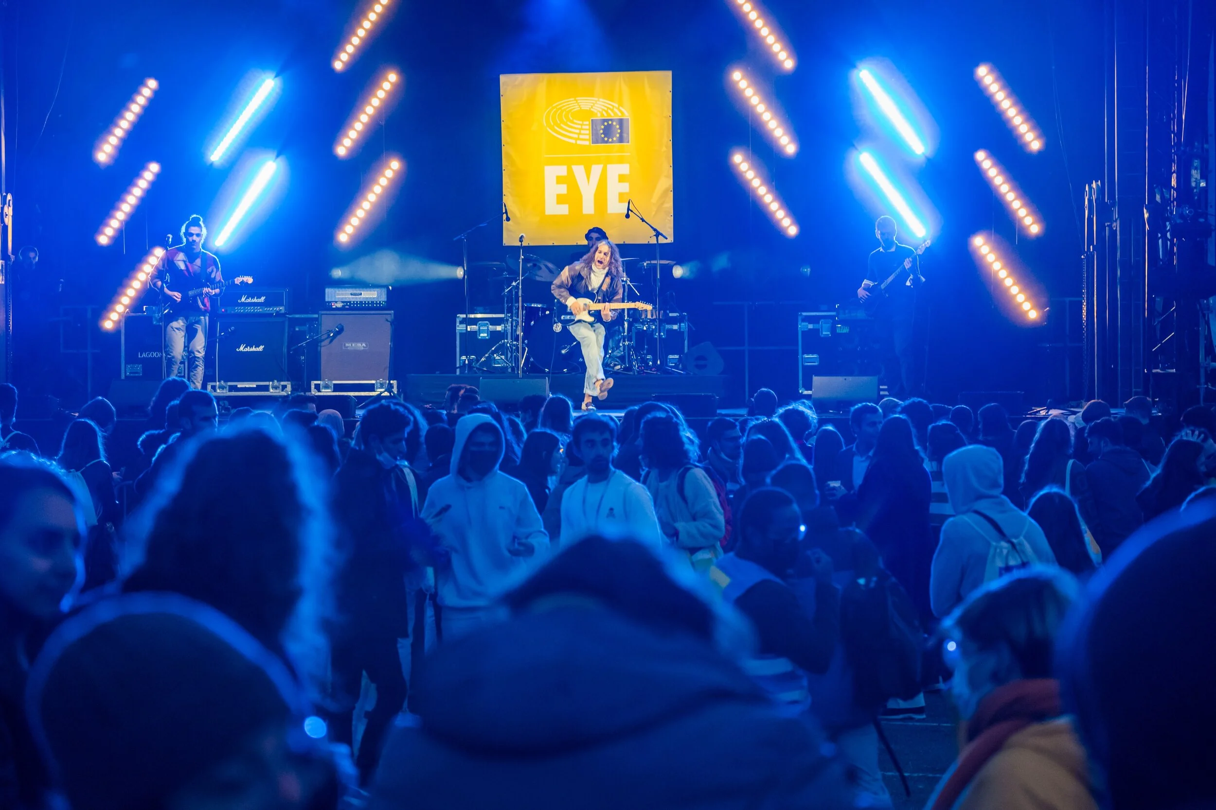 The audience watches a performance on a stage lit in blue, with beams of light and a yellow screen in the background.