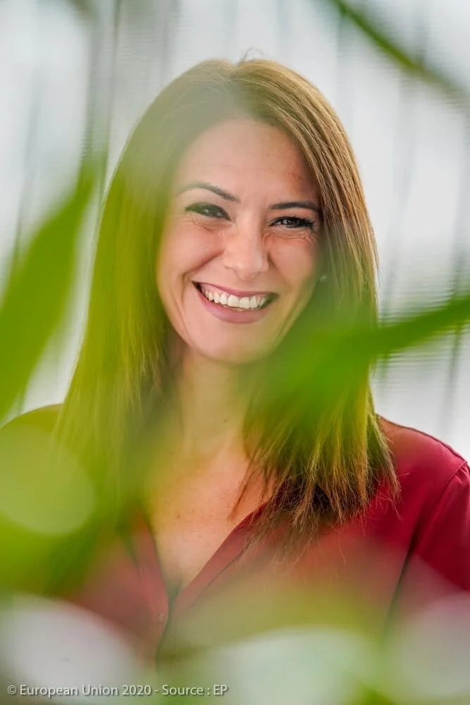 A portrait of a smiling woman with long hair, with blurred green leaves in the foreground.