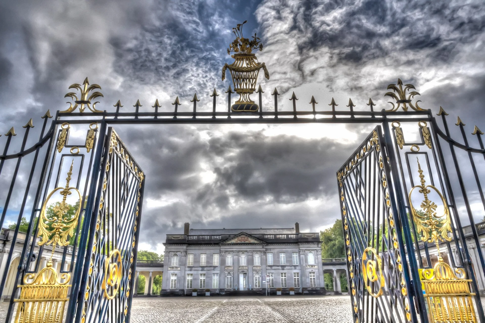 Le château de Seneffe vu à travers son portail monumental en fer forgé et dorures, sous un ciel nuageux.