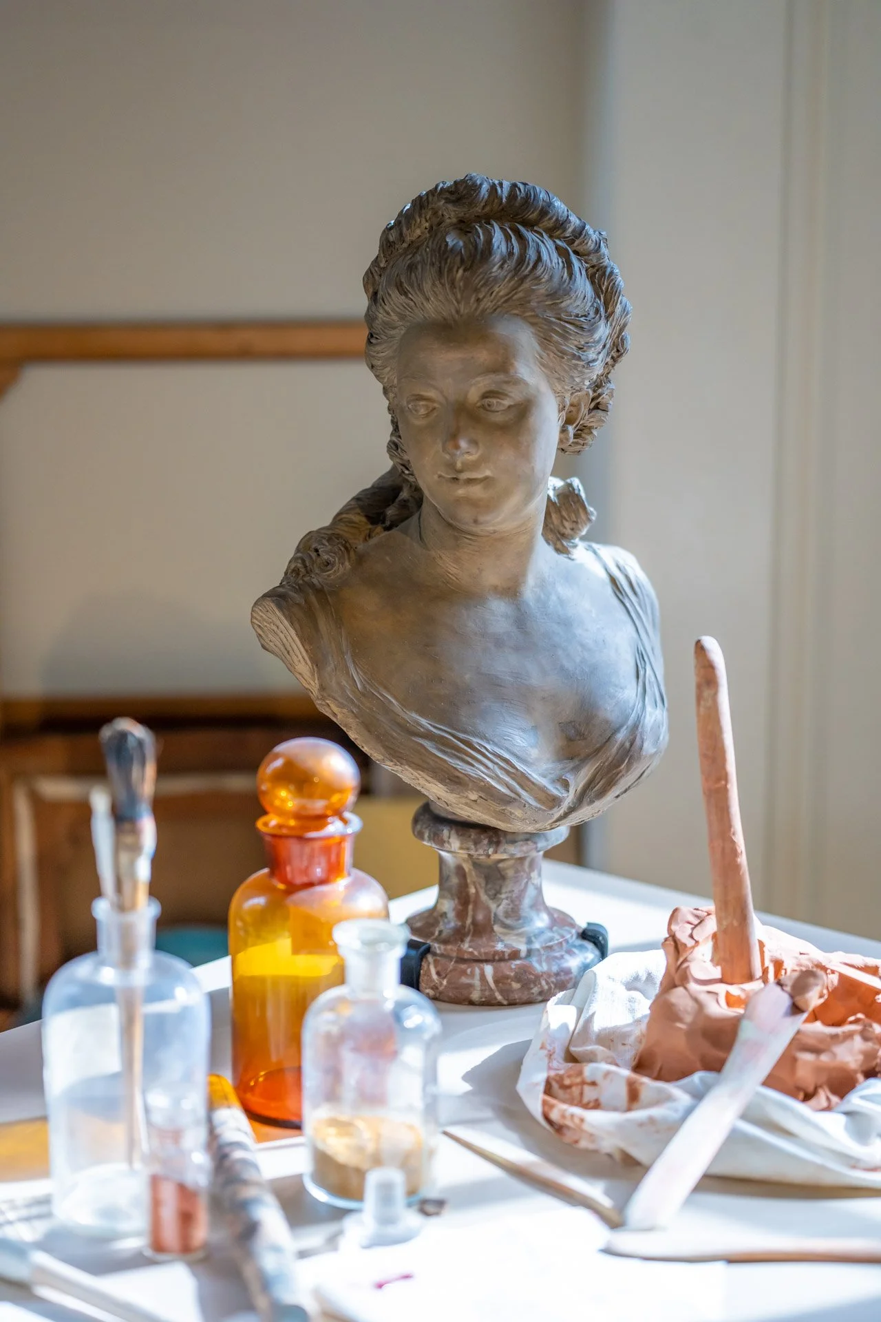 Buste de femme en pierre, placé sur une table avec des bouteilles en verre, des outils et de la pâte à modeler.