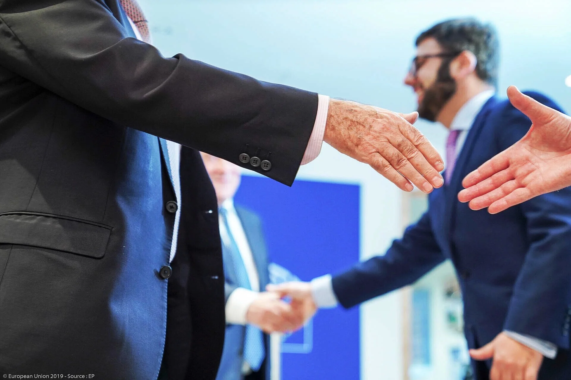 Participants in suits greet one another at a business meeting, with outstretched hands in the foreground.