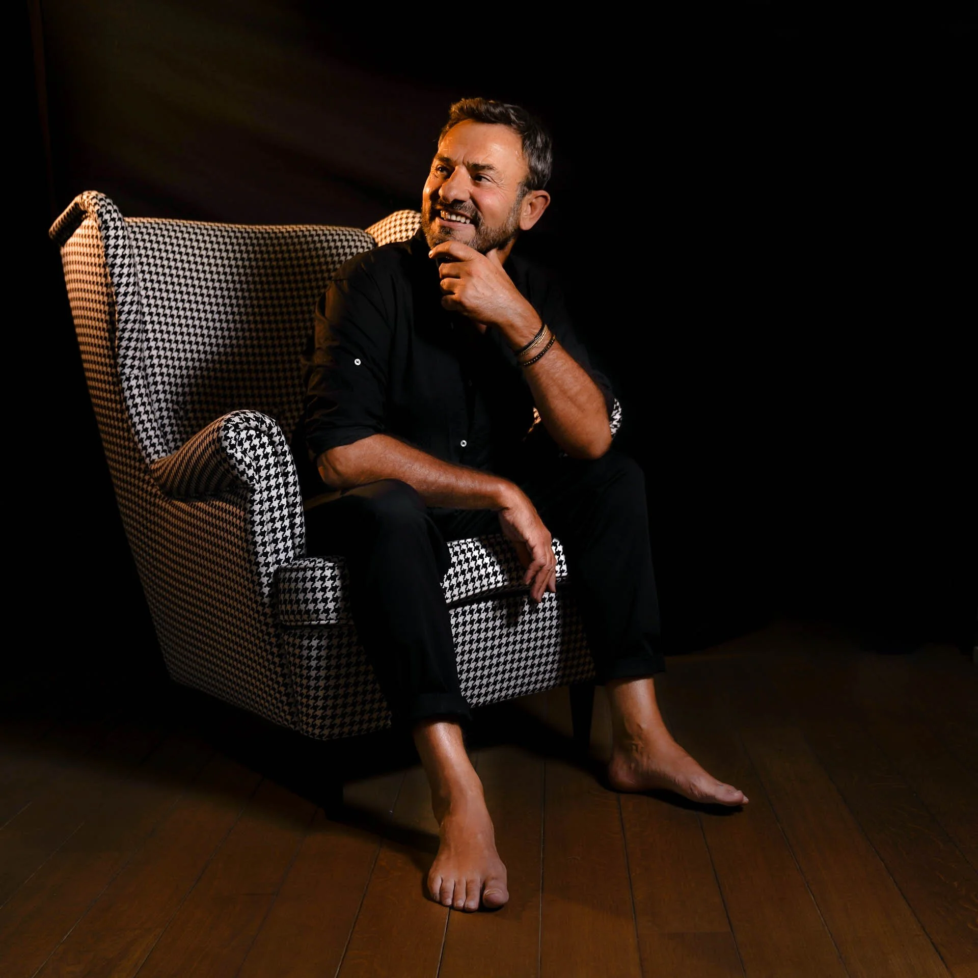 Homme assis sur une chaise à motif pied-de-poul, cheveux foncés, barbe, vêtements noirs, expression pensante, environnement sombre.