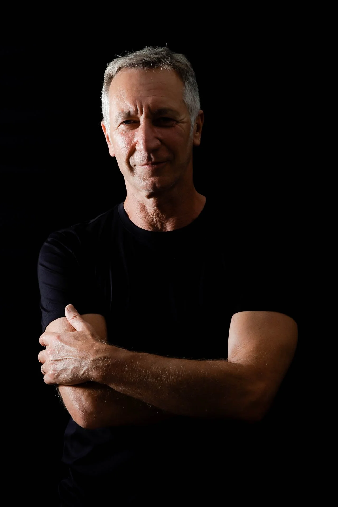 Un homme avec des cheveux gris, portant un t-shirt noir, qui se tient contre un fond noir, croisant les bras, regardant légèrement à gauche, avec une expression confiante.