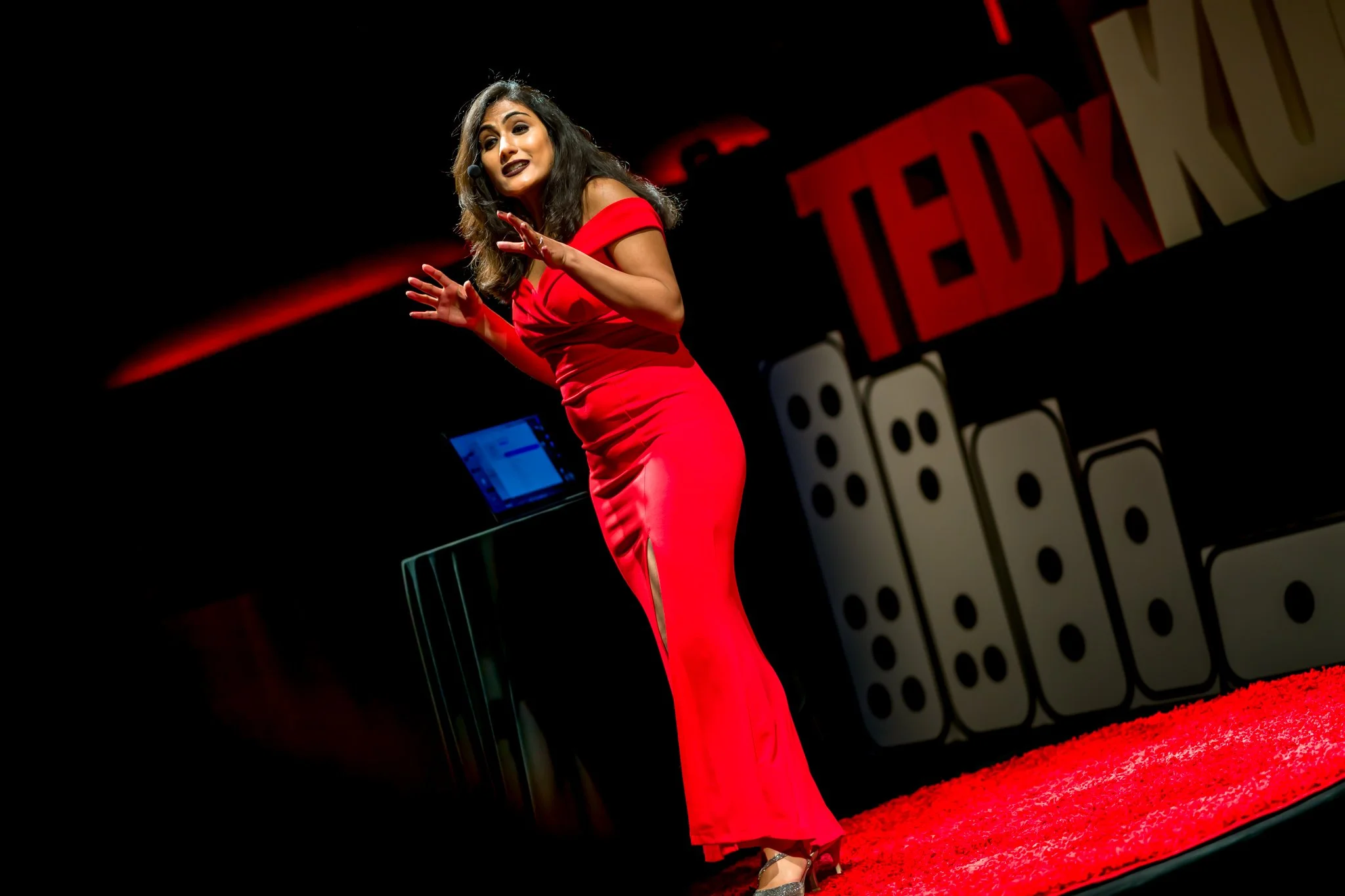 Femme en robe rouge parlant lors d'une conférence TEDx, scène avec logo TEDx et décor de dominos.