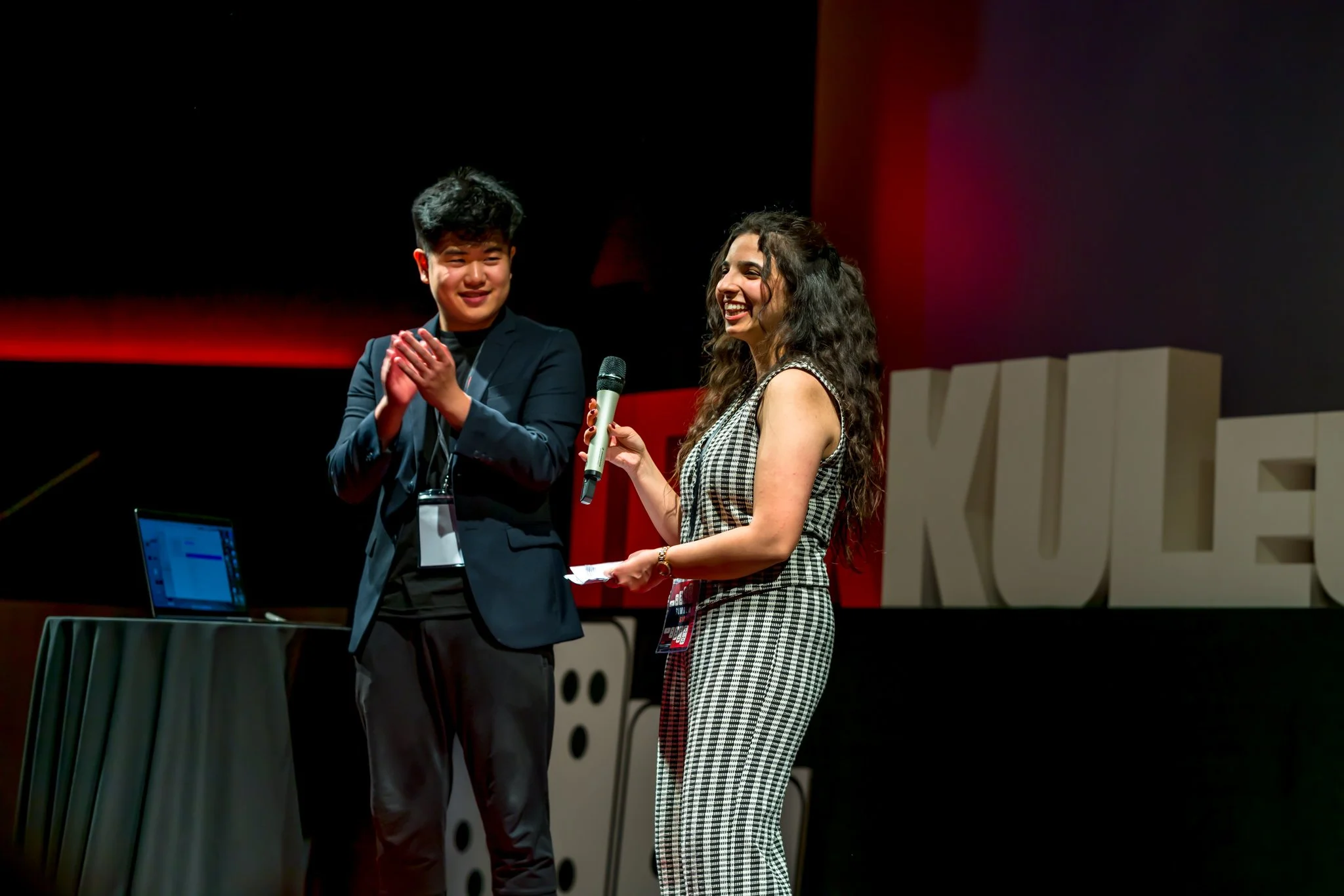 Photographie professionnelle pendant une conférence TEDx. Un homme applaudit tandis qu'une femme tient un microphone sur scène, avec un ordinateur portable posé sur une table à gauche.