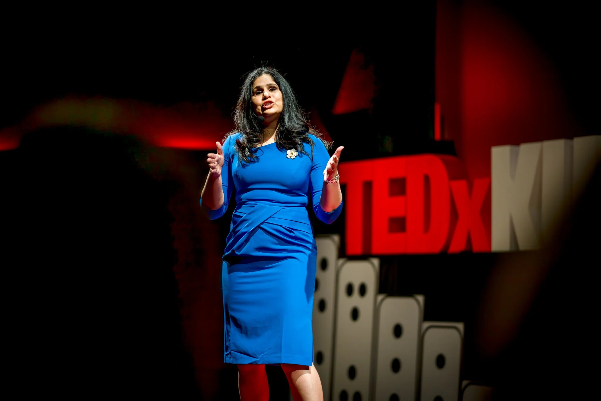 A woman in a blue dress speaks on a dark stage beside large red TEDx letters.