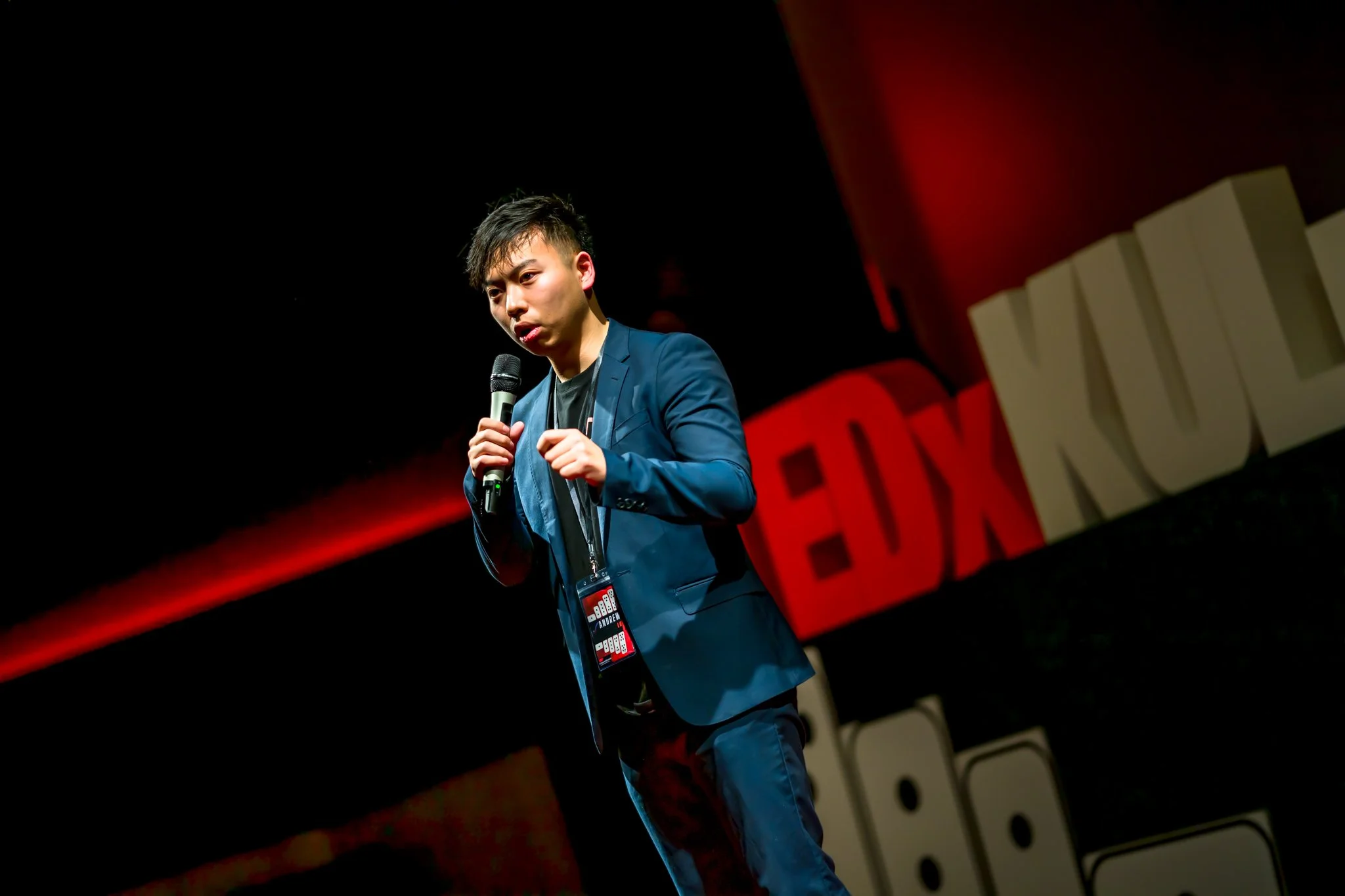 A man in a blue suit holding a microphone on stage at a TEDx event, with the TEDx logo in the background.