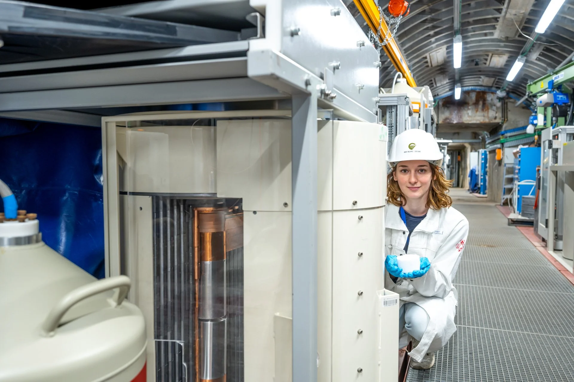 Photographie d'entreprise d'une ingénieure en combinaison de sécurité et casque blanc, travaillant dans une installation industrielle avec de l'équipement technique.
