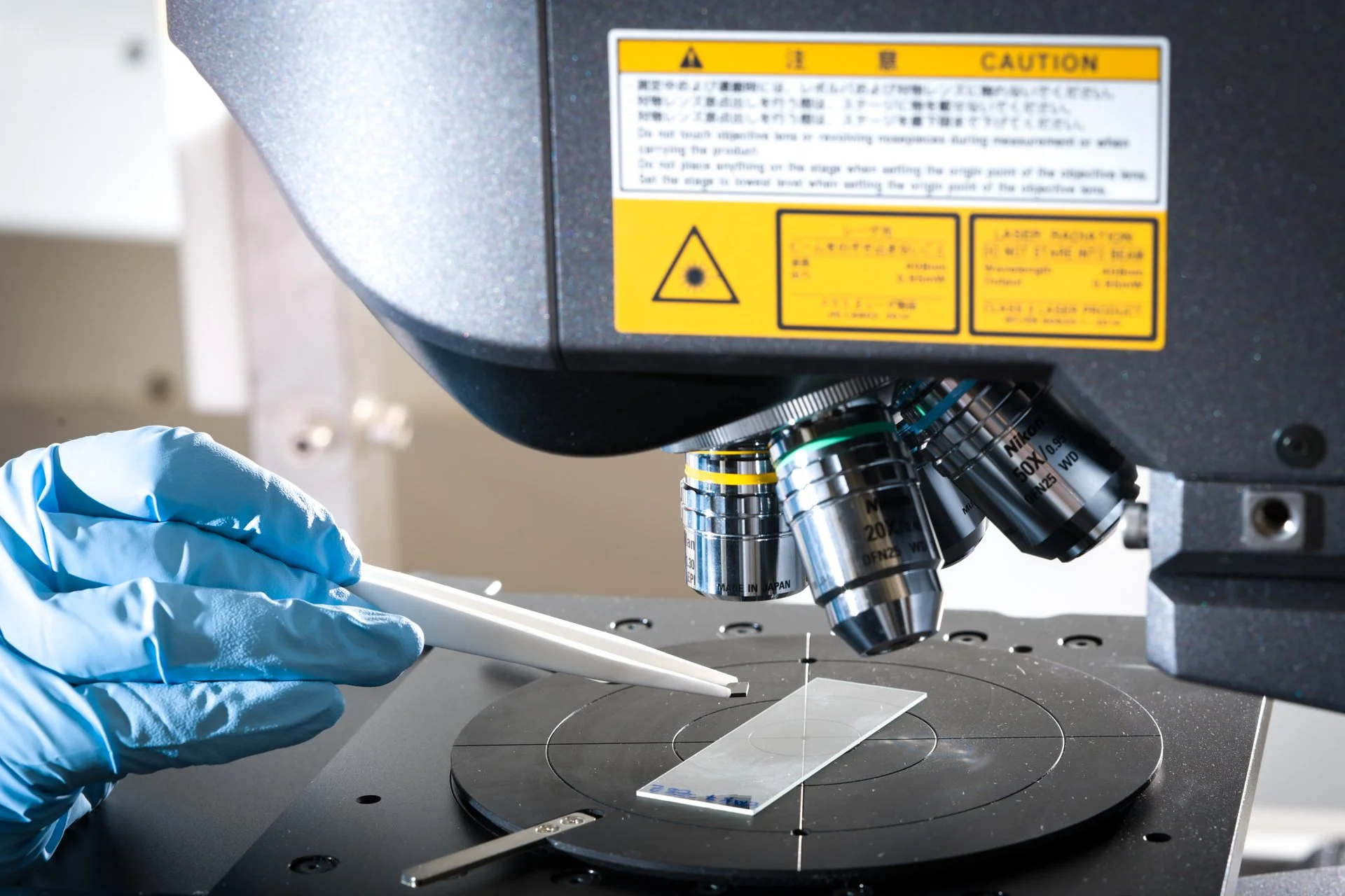 A person wearing blue gloves is using tweezers to handle a small object under a microscope in a science laboratory.