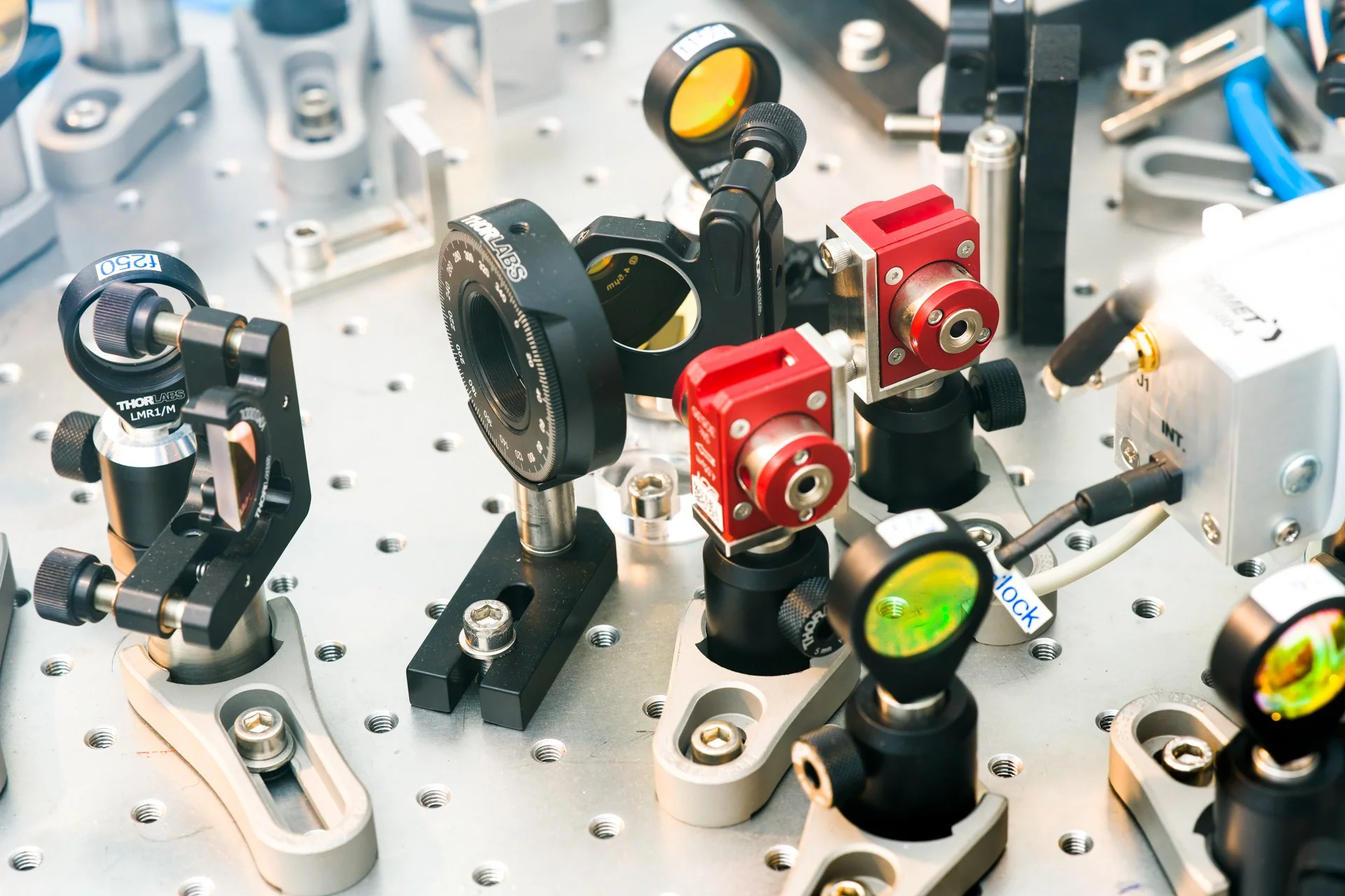Photo d'entreprise qui montre les composants optiques et instruments sur une table optique dans un laboratoire, comprenant des miroirs, des supports de montage et des dispositifs de réglage.