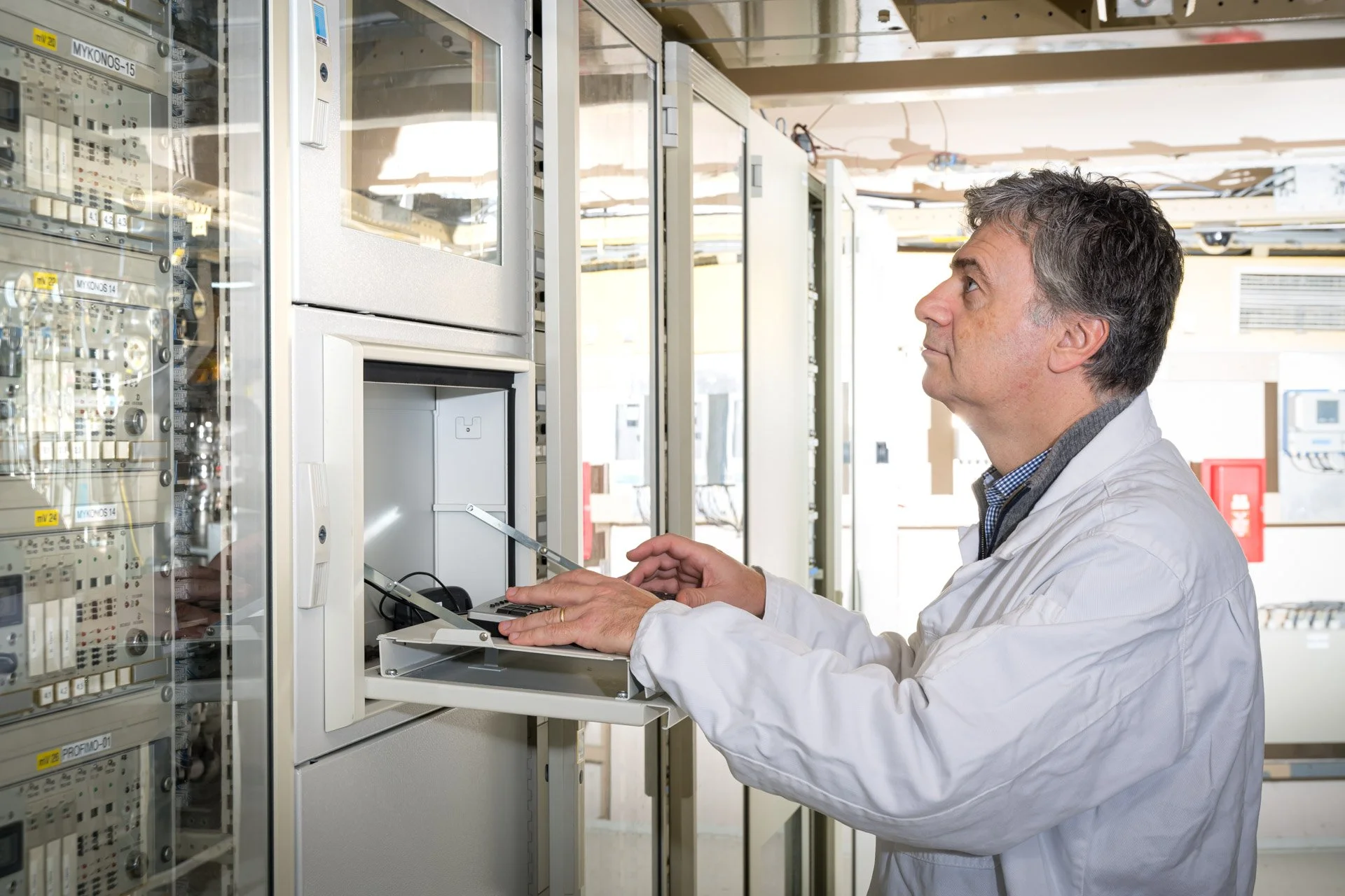 A man in a lab coat working in a technical room or a control centre with electronic equipment.