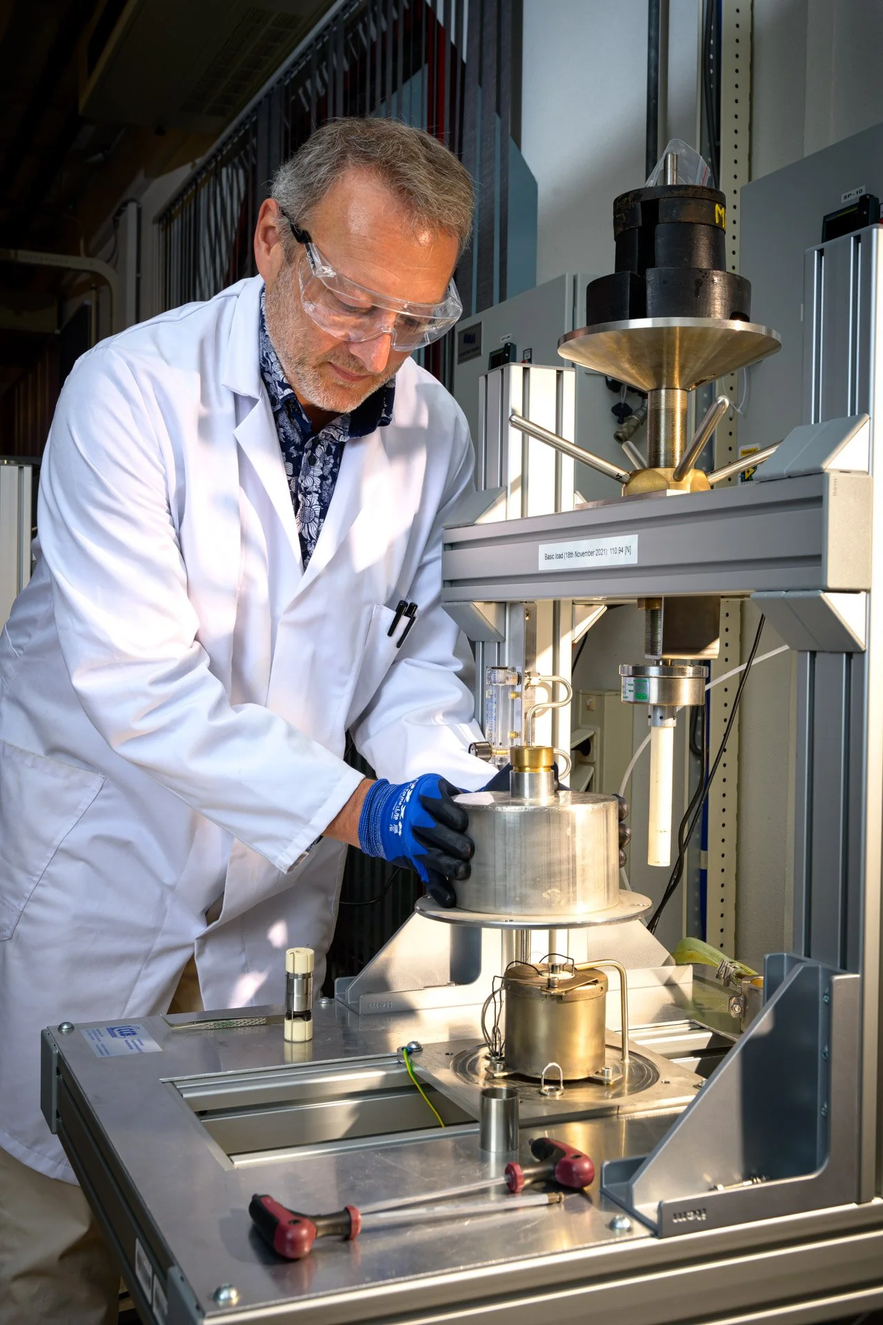 A scientist in a white lab coat is carrying out an experiment using complex laboratory equipment, whilst wearing safety goggles and gloves, in an industrial laboratory.