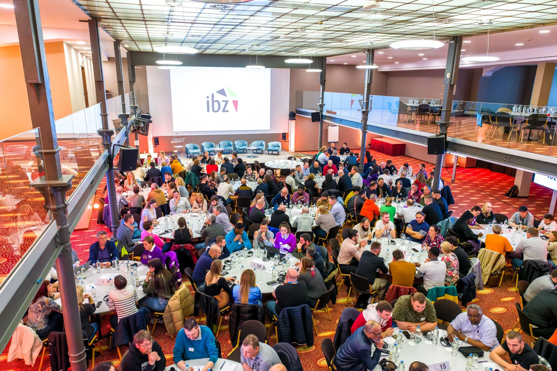 Grand événement photographié par ISOWAY. Salle de conférence avec de nombreuses personnes assises autour de tables rondes, projetant un écran affichant le logo "ibz".
