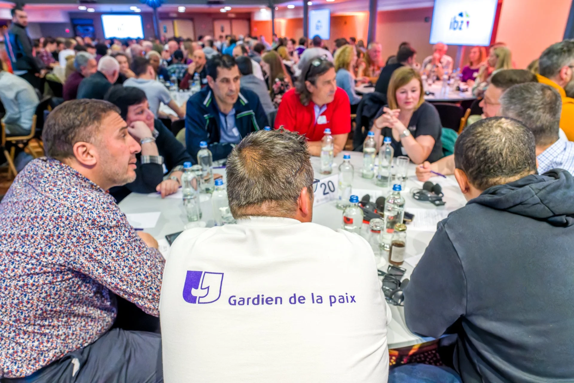 Une conférence débat photographiée par ISOWAY. Un groupe de personnes assises autour d'une table lors d'une réunion ou conférence, avec des bouteilles d'eau et des microphones. Une personne porte un t-shirt avec l'inscription 'Gardien de la paix'. Be
