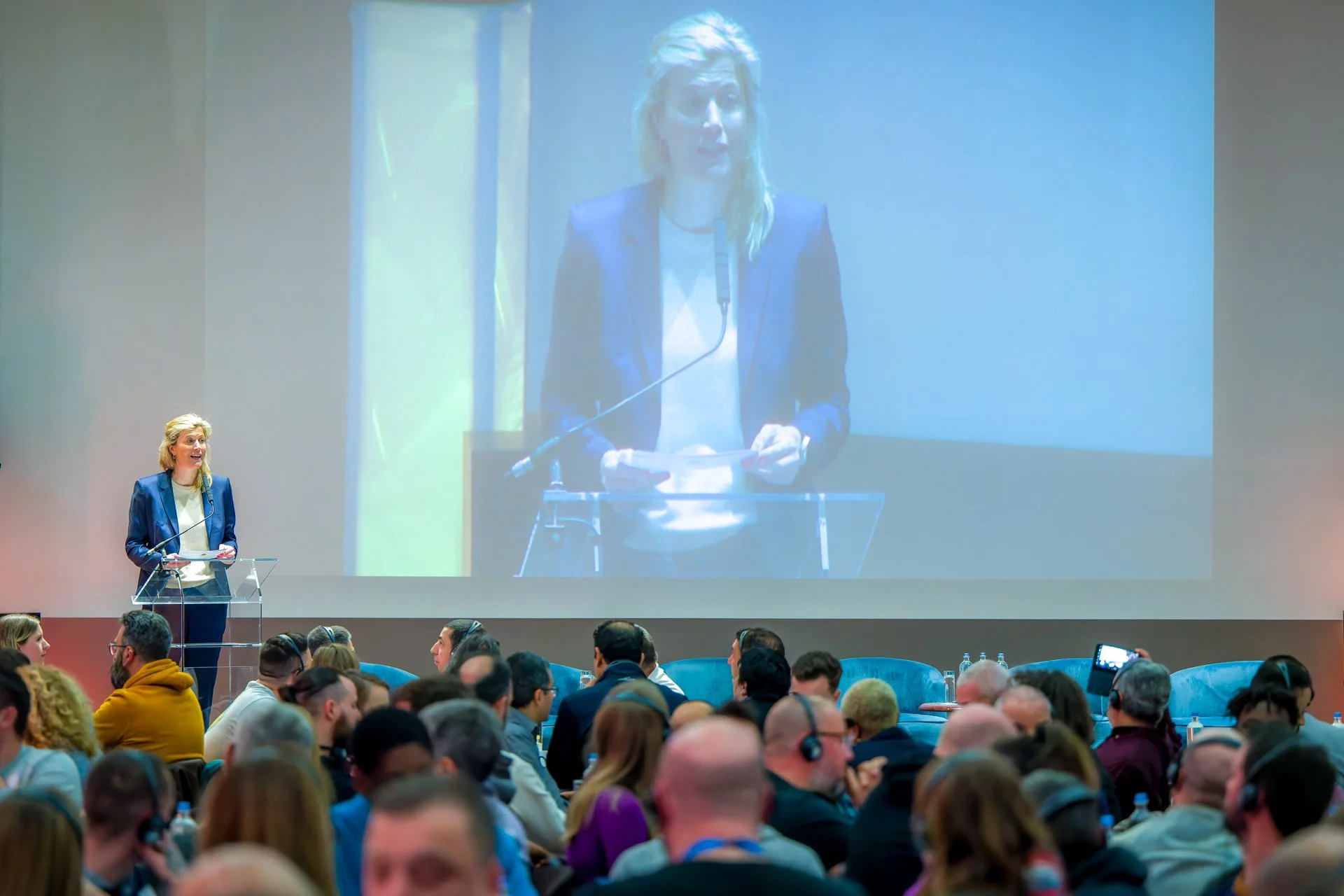 Photo d'une conférence où l'on voit une personnalité prendre la parole. Une femme en costume parle à une conférence, debout à un pupitre avec un microphone. Elle est projetée sur un grand écran derrière elle. Un public de personnes assises écoute att