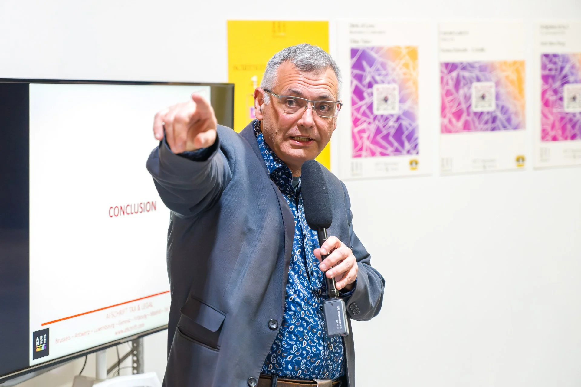 Vincent Parissis s’exprime au microphone et pointe vers le public devant des œuvres exposées à la BXL ART NFT Gallery.