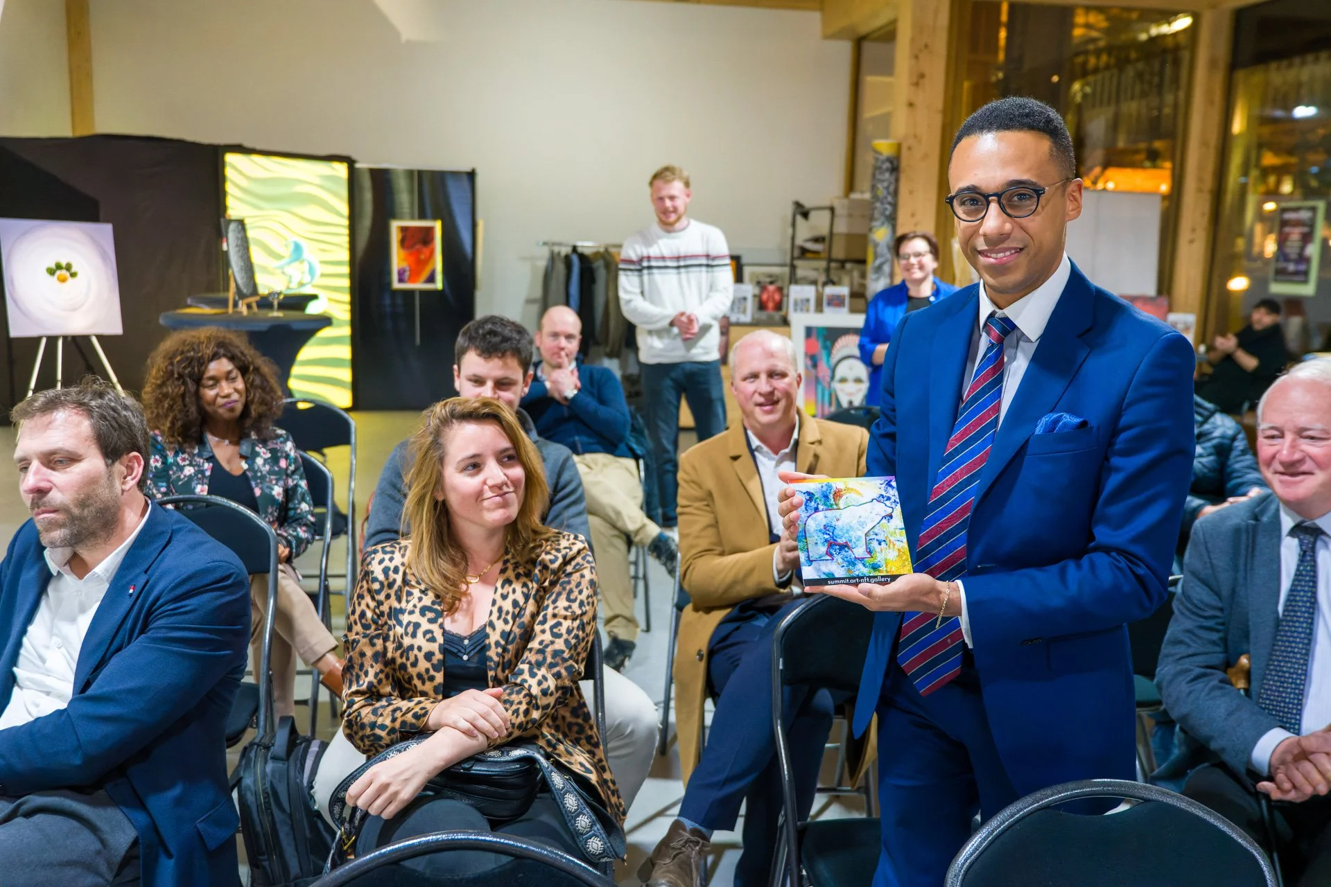 A group of people sitting in a room, with a man in a blue suit holding a colourful work of art. The participants are looking intently ahead. Paintings are on display in the background on easels.