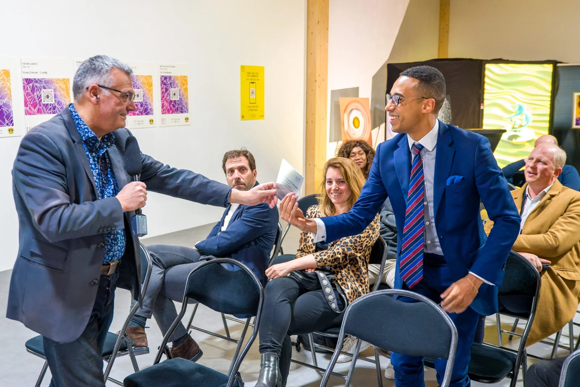 Two smartly dressed men are exchanging a document in a conference room, whilst spectators sit and watch them.