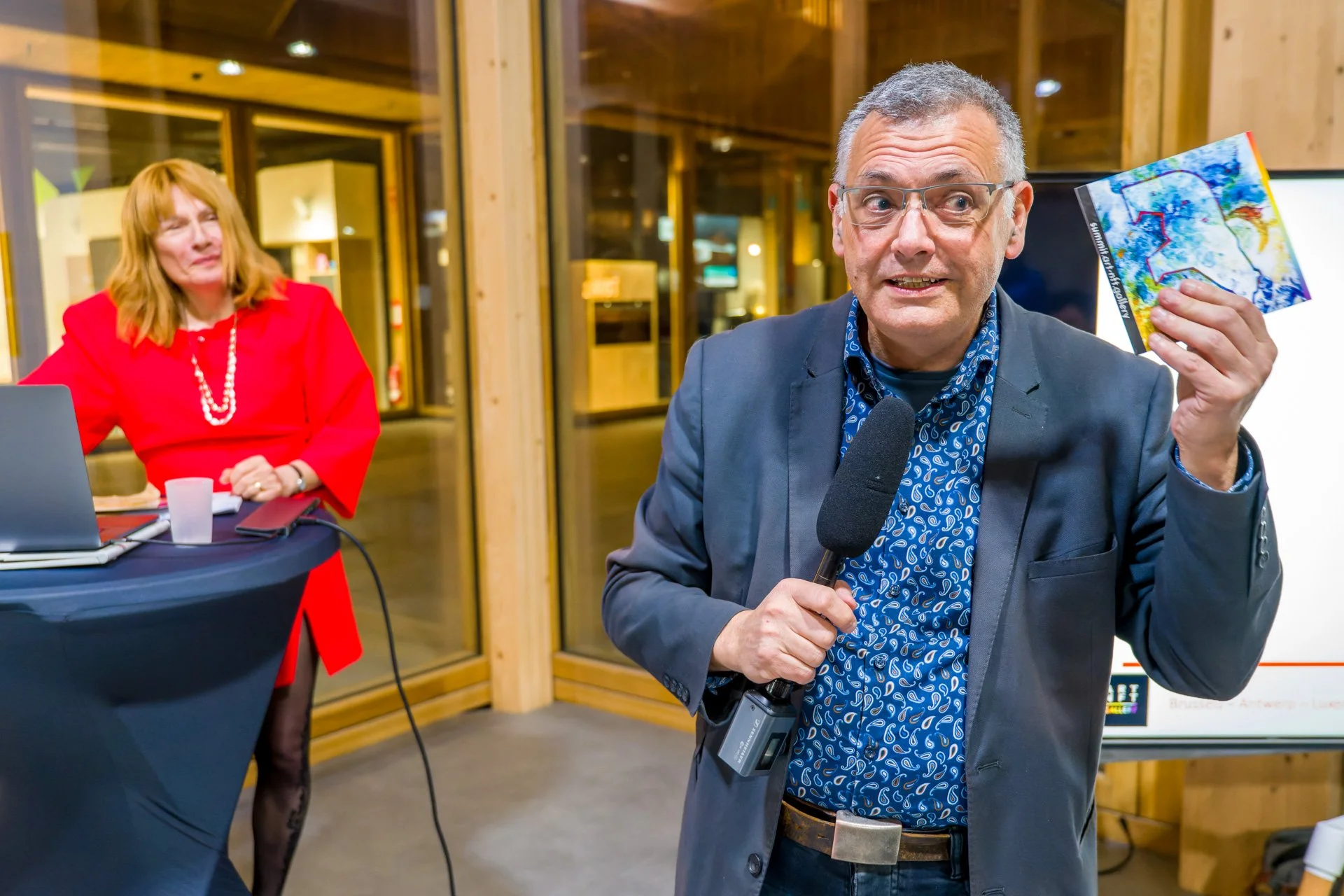 Vincent Parissis tient un microphone et présente une illustration bleue et jaune, avec Typhanie Afschrift à l’arrière-plan.