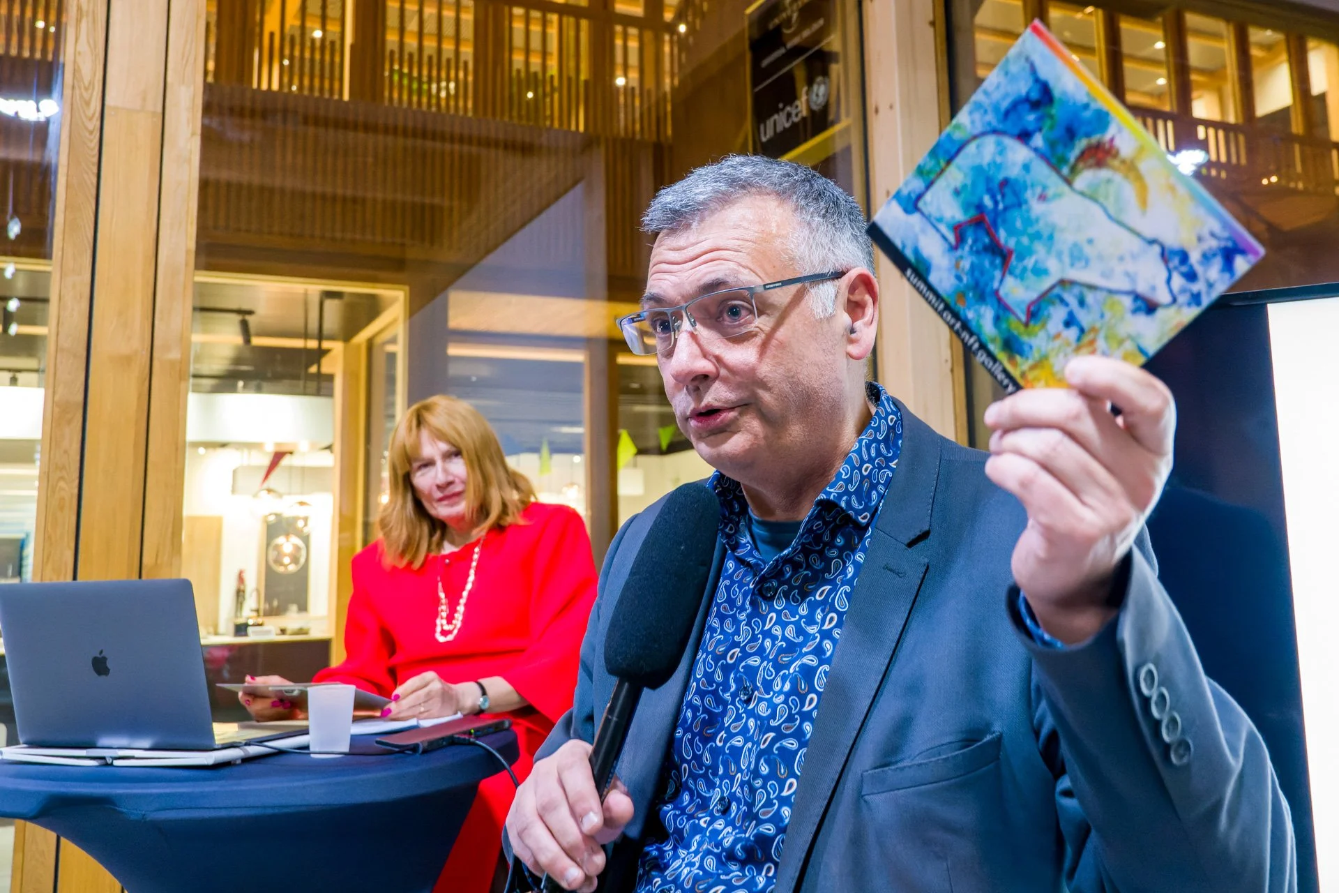 Vincent Parissis montre une illustration bleue et jaune tout en tenant un microphone, avec Typhanie Afschrift à l’arrière-plan.
