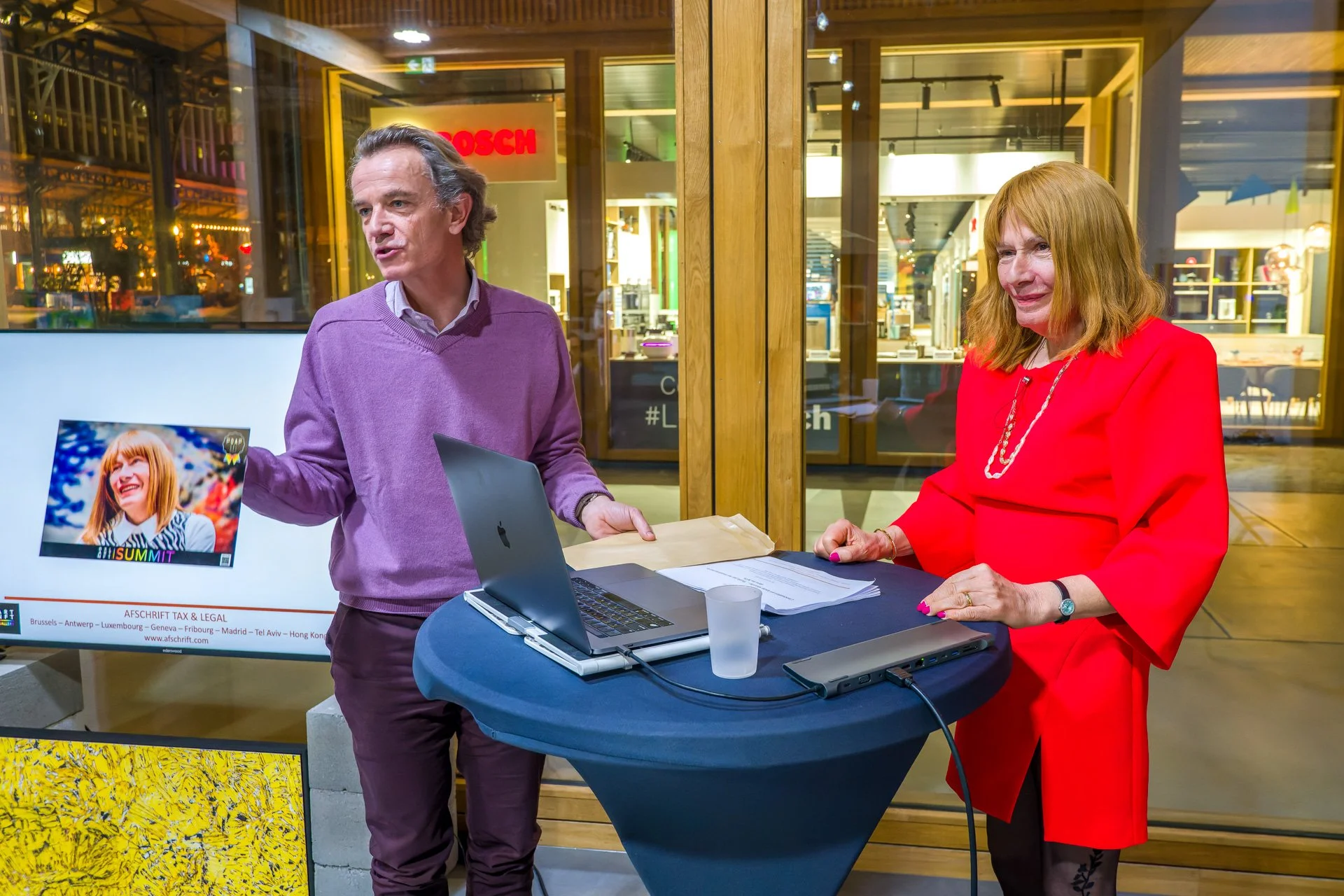 Two people are chatting next to a podium; the man is holding a photograph and the woman is dressed in red, against a backdrop of a modern urban setting with shop fronts.