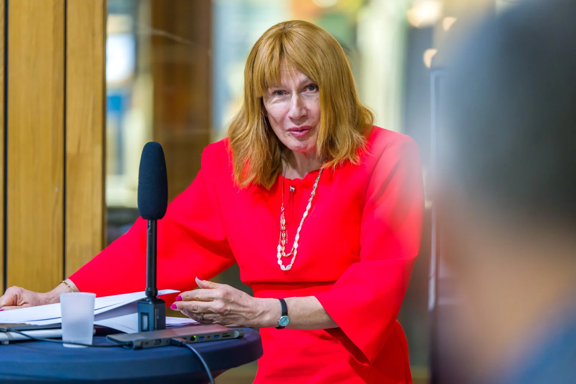 Typhanie Afschrift is speaking into a microphone and a laptop, with a blurred participant in the foreground.