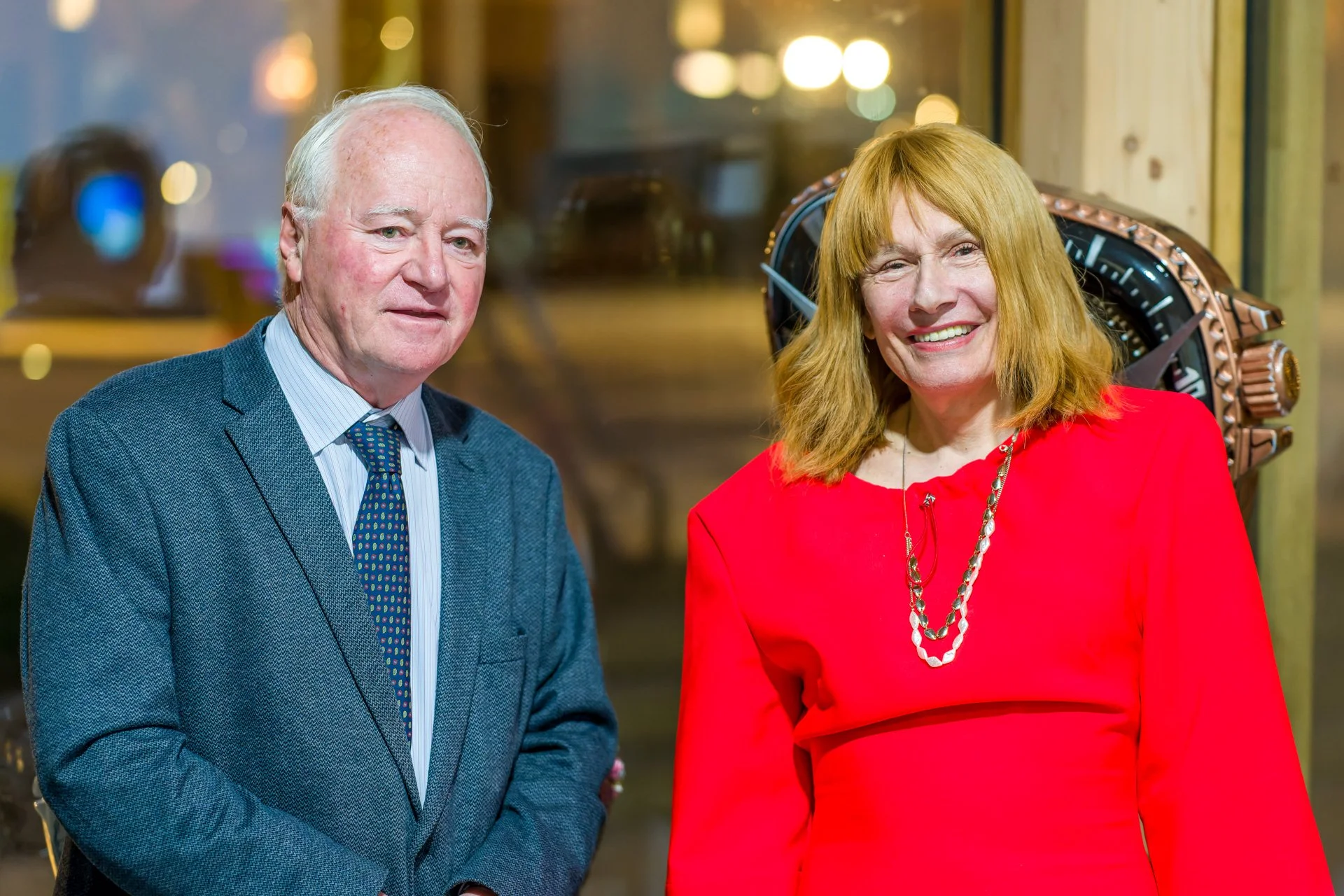 Two people smiling – a man in a blue suit and a woman in a red dress – standing in front of a shop window with lights in the background.